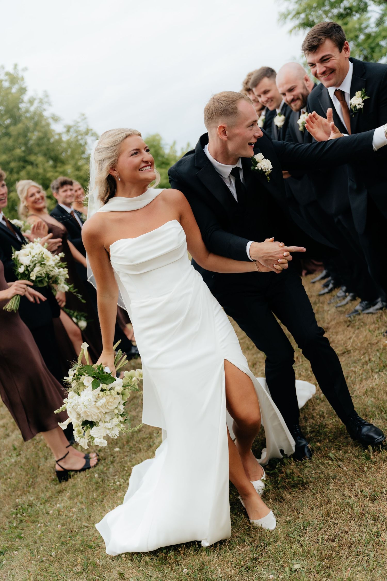 Bride in white strapless gown holding bouquet walks with groom in black suit, surrounded by cheering wedding party outdoors.