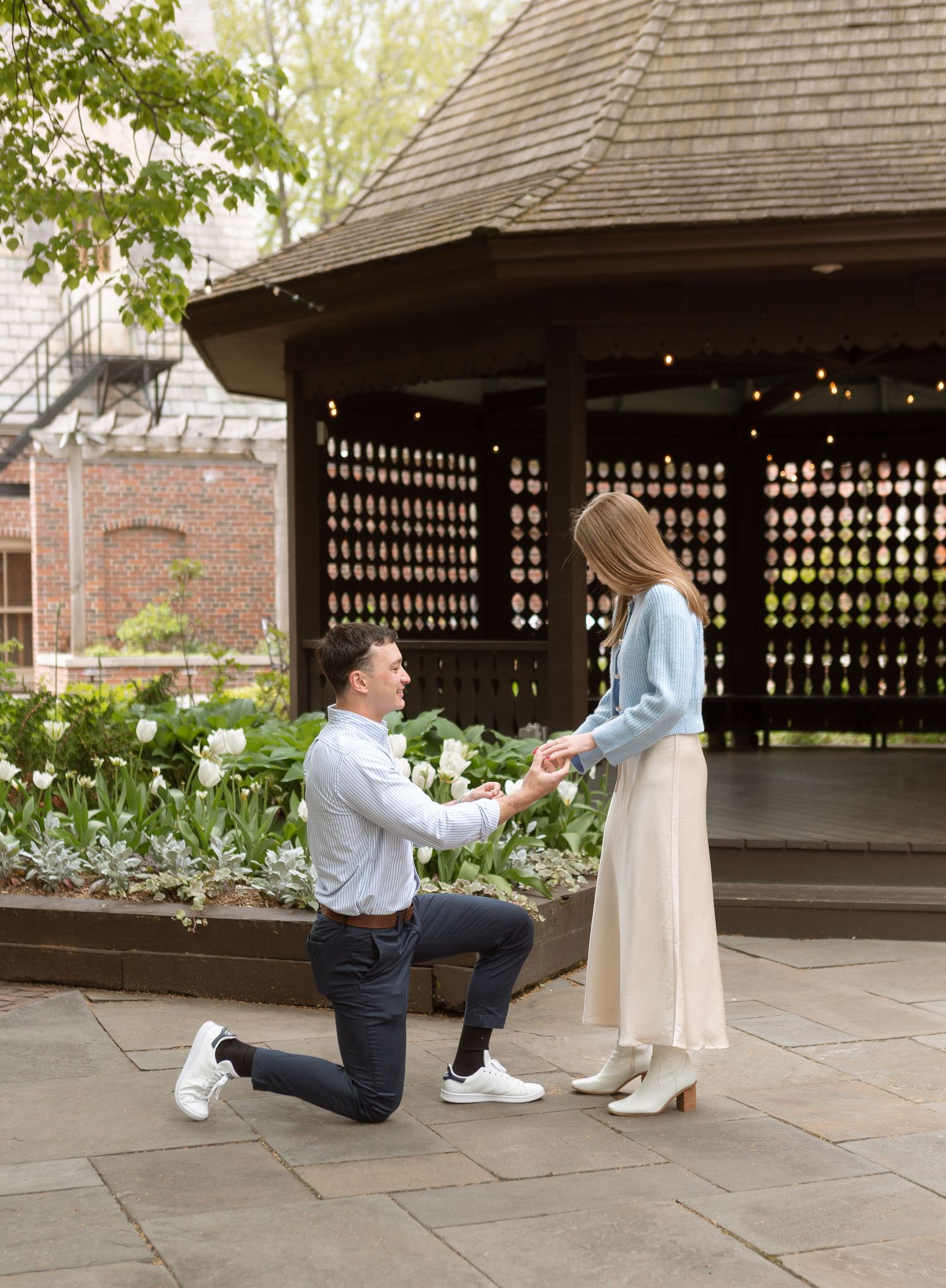 Man proposes on one knee to woman in front of a garden gazebo with white tulips in bloom.
