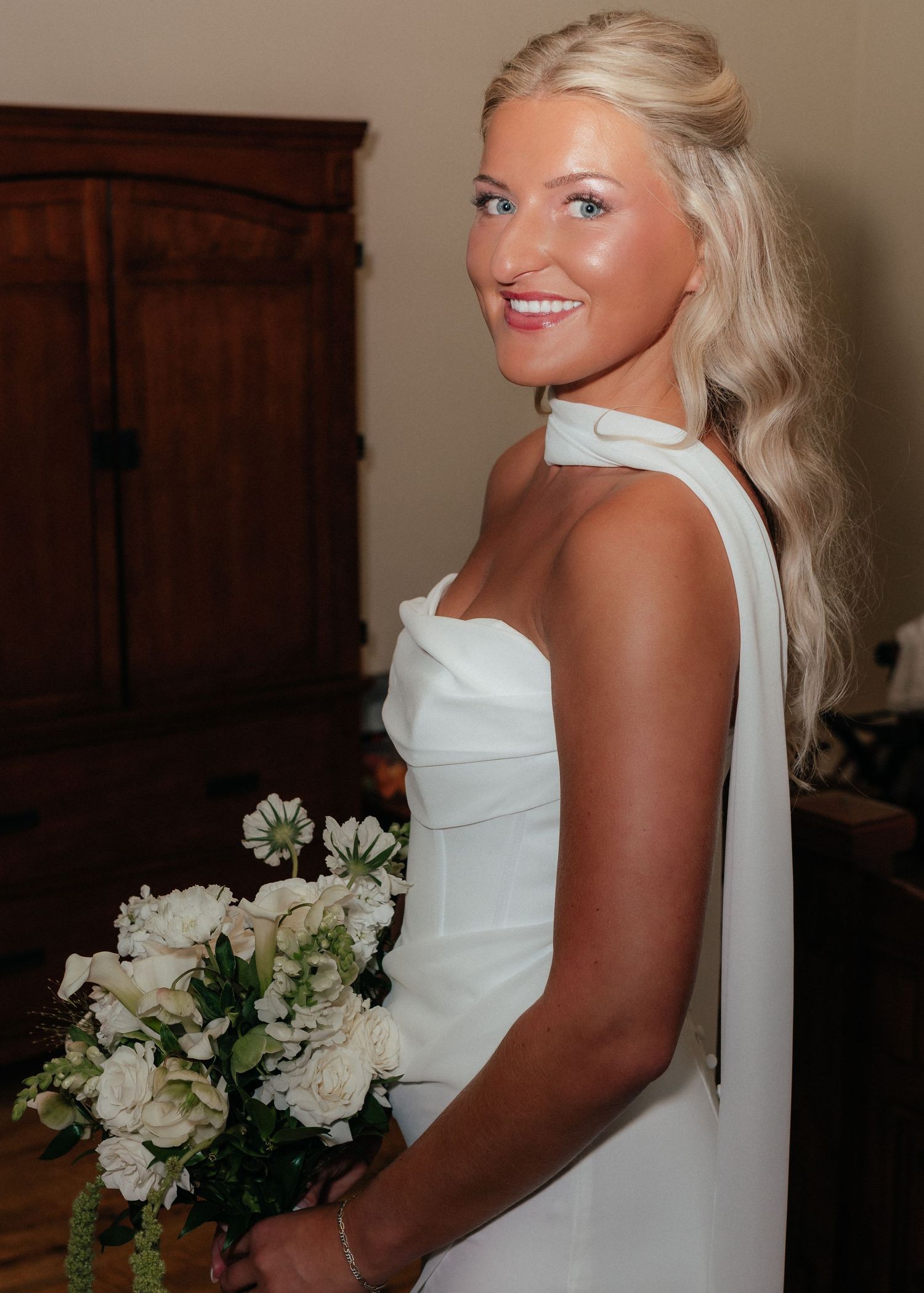 Elegant bride in white halter gown holds bouquet of white roses, smiling over her shoulder.