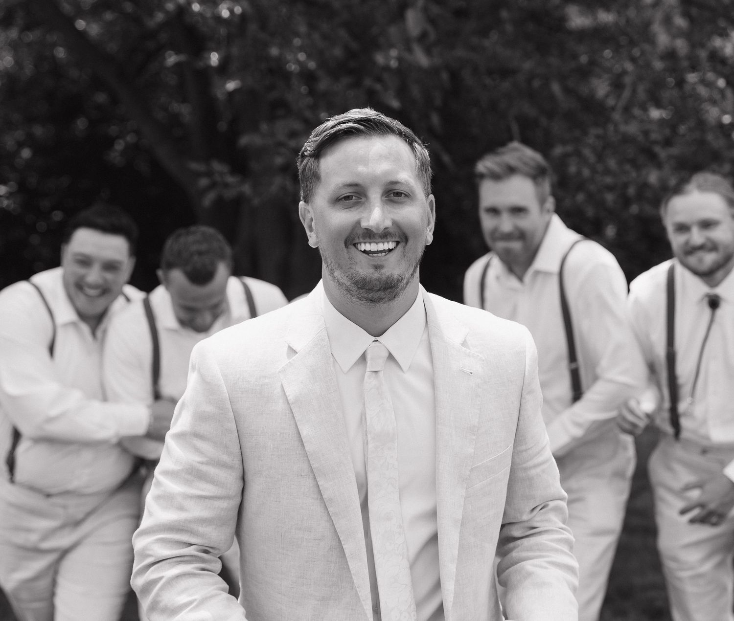 A black and white photograph of a smiling man in a suit standing outdoors with several other men in the background.