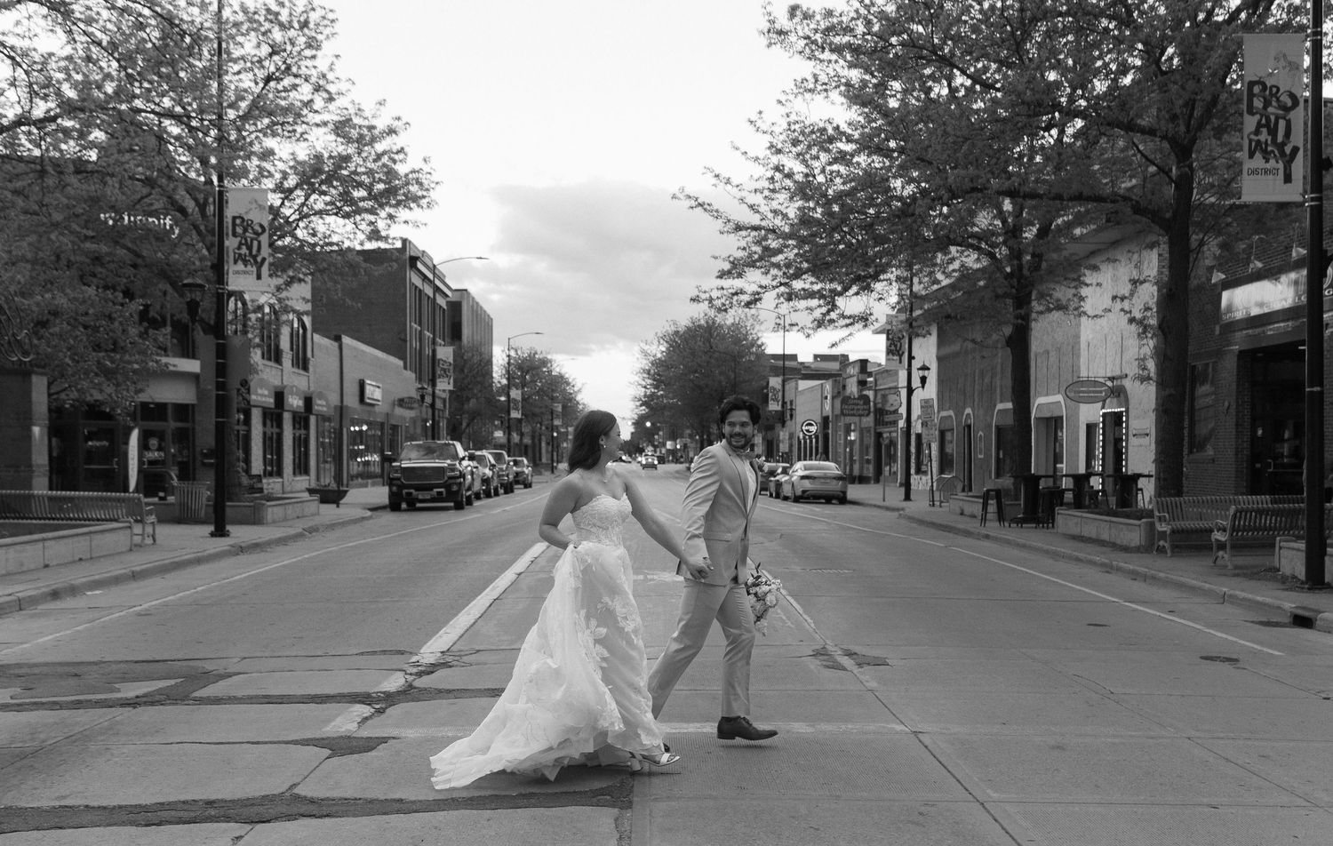 A black and white image of a person wearing a wedding dress walking down a city street lined with trees and buildings.