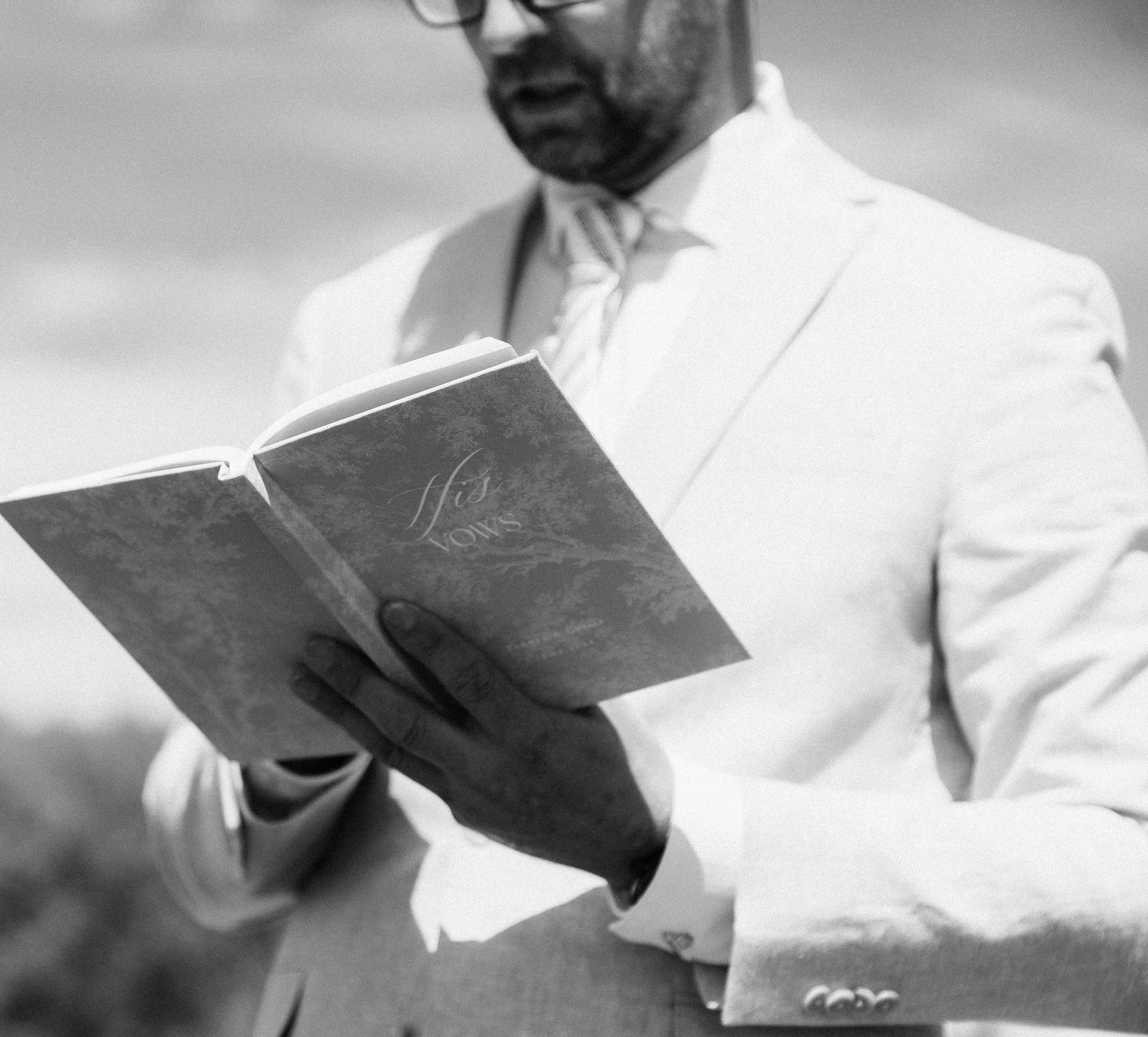 Man in white suit reading from a book outdoors in black and white photo.