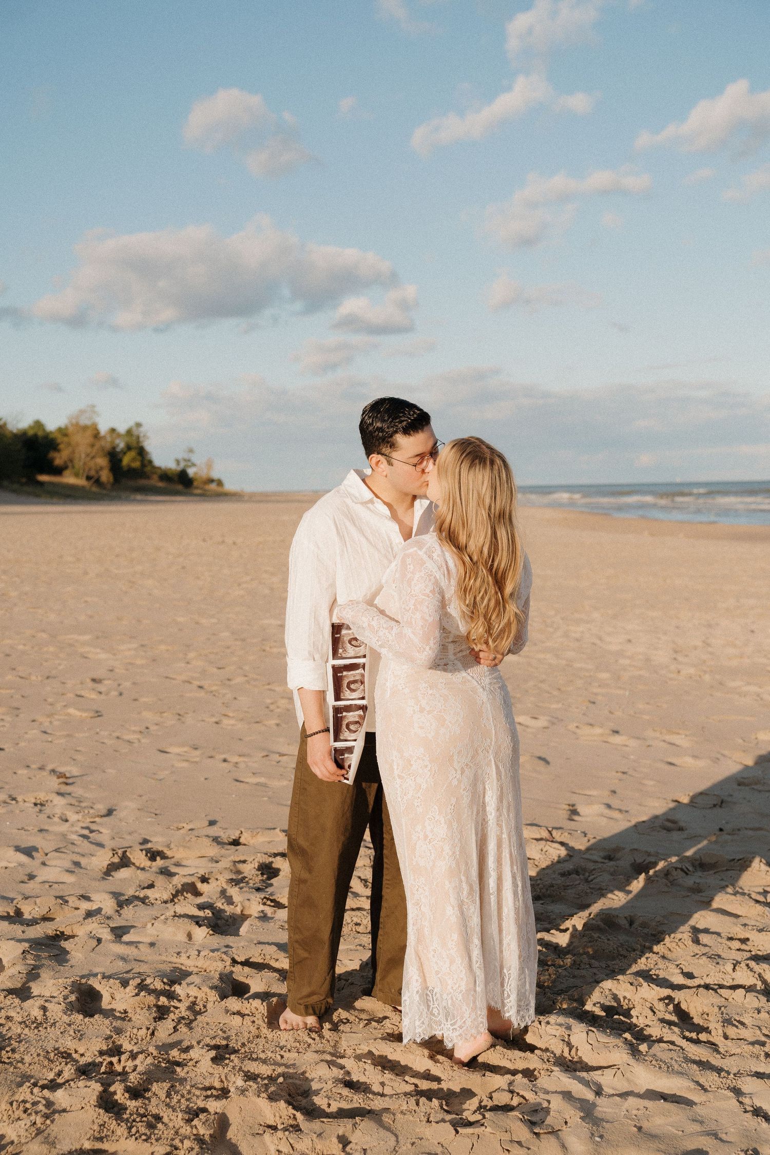 Couple kissing on sandy beach holding ultrasound photos, woman in lace dress, pregnancy announcement at sunset.