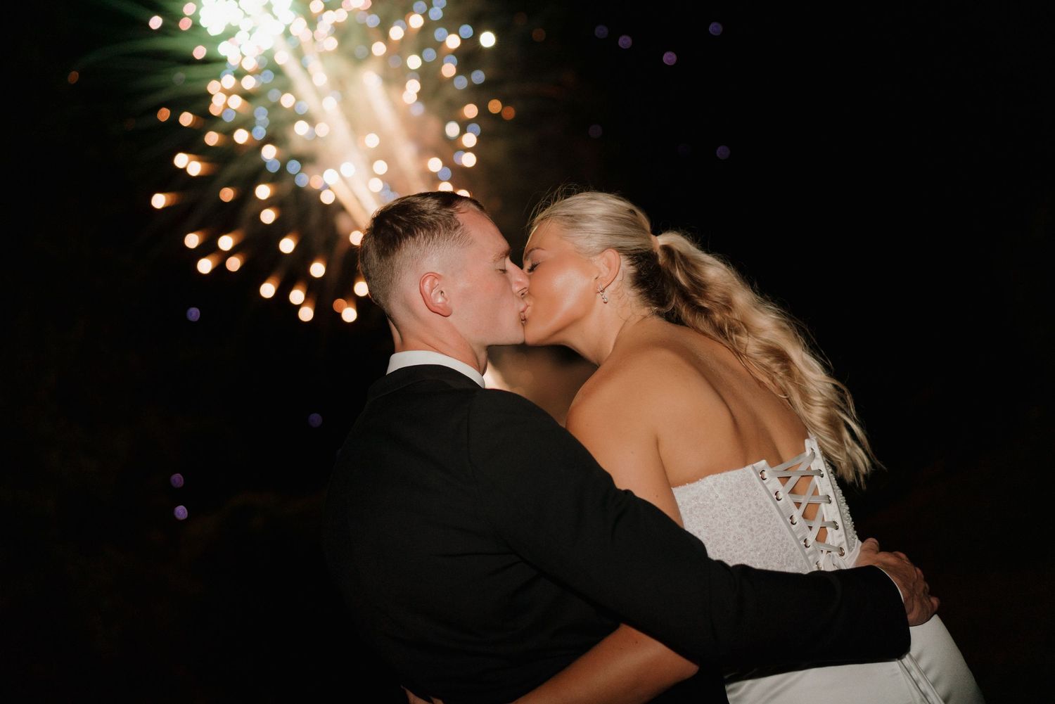 Newlywed couple sharing a romantic kiss in front of dazzling fireworks display at their wedding reception.