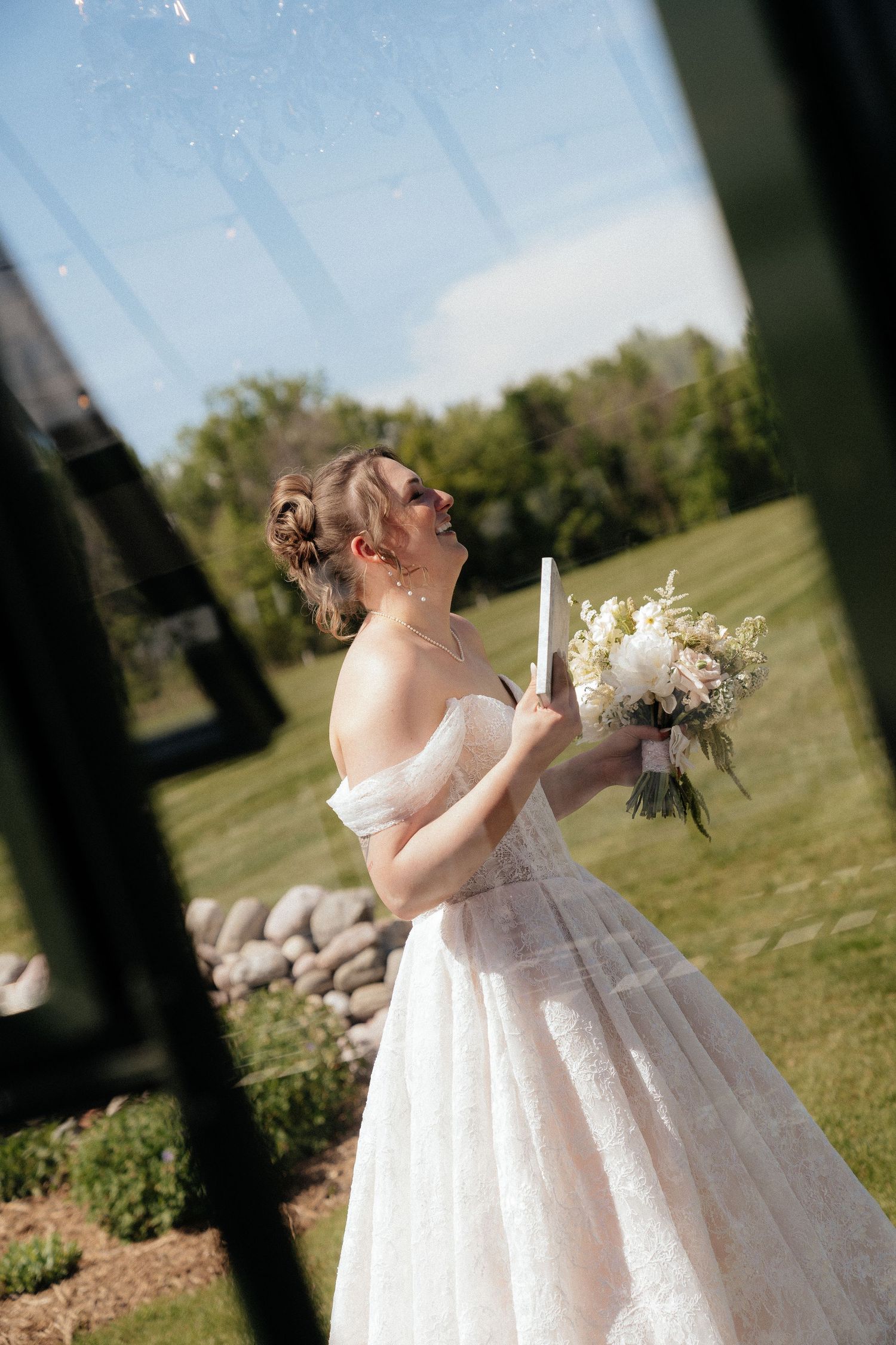 Smiling bride in off-shoulder white gown holds bouquet and candle outdoors on sunny day.