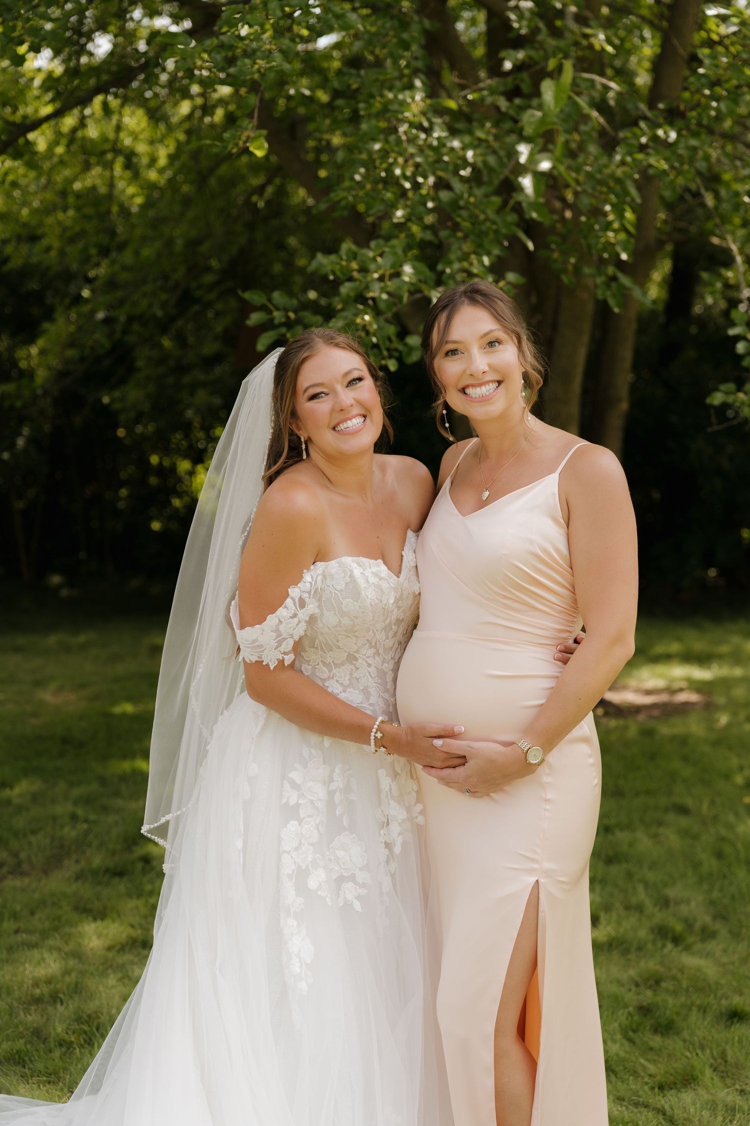 Two women, one in a white wedding dress and the other in a pink dress, stand together in a garden setting.
