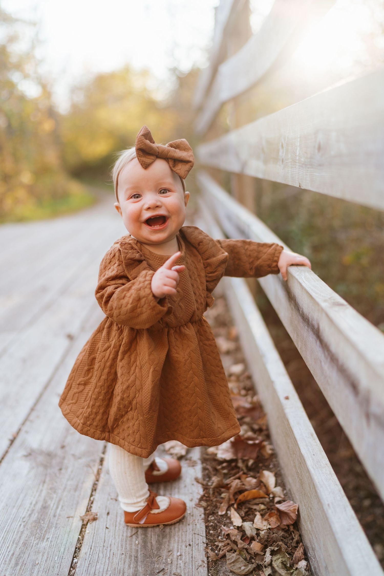Smiling baby girl in brown dress and bow headband standing on wooden bridge surrounded by autumn leaves.