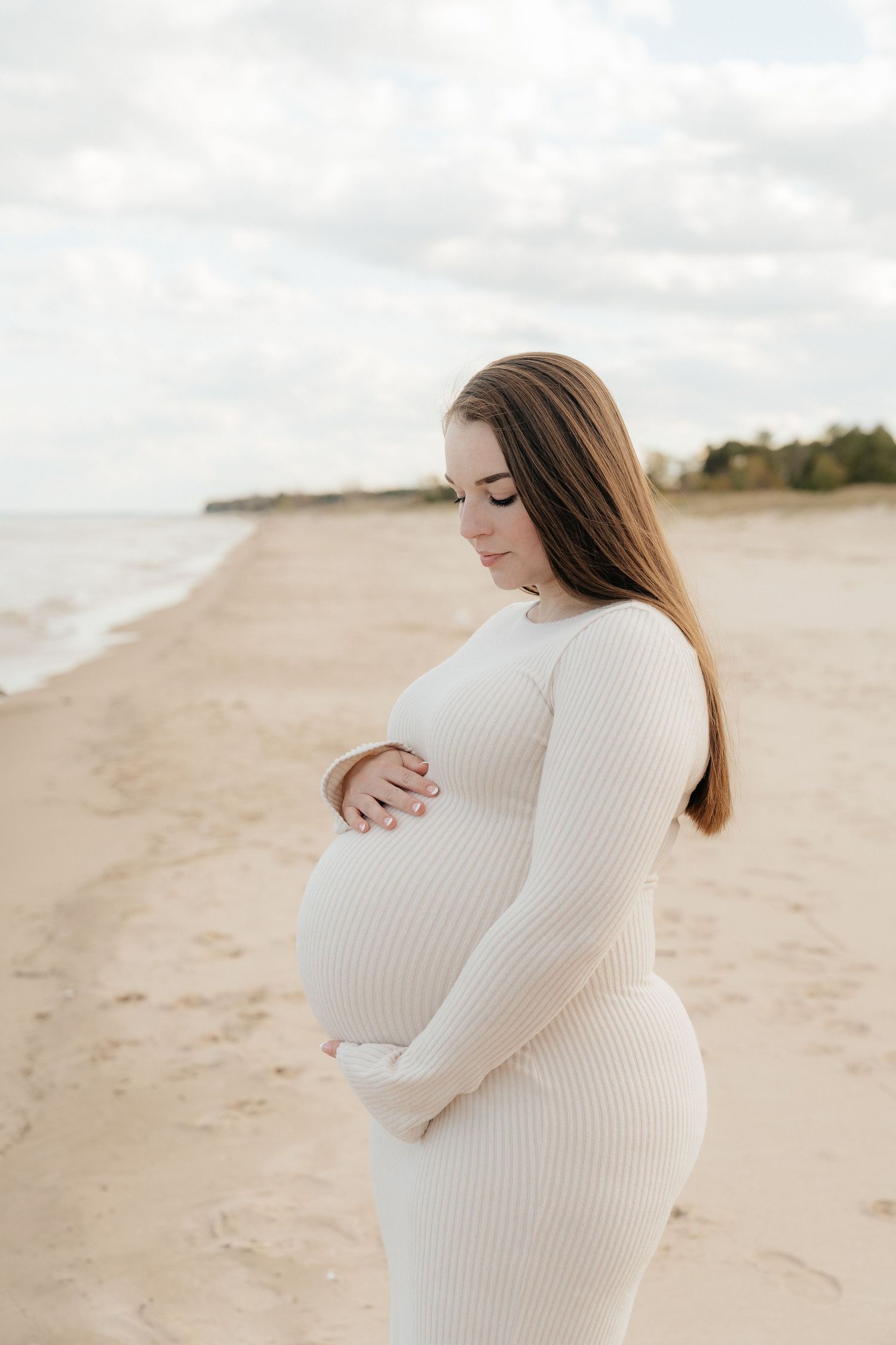 A pregnant woman in a white ribbed dress cradles her baby bump while standing on a serene sandy beach.