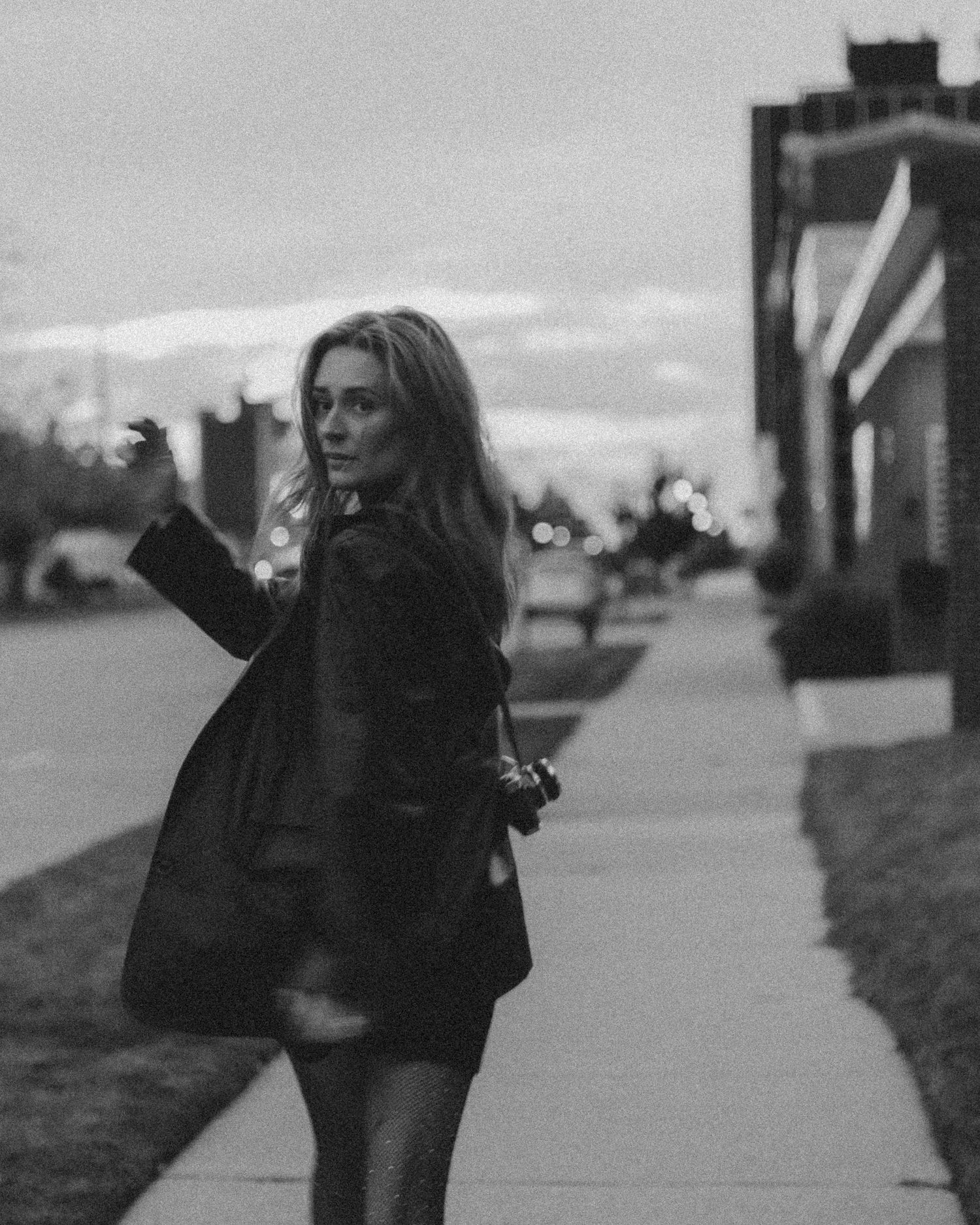 Black and white photo of a woman in a dark coat walking on a sidewalk at dusk, glancing back over her shoulder.
