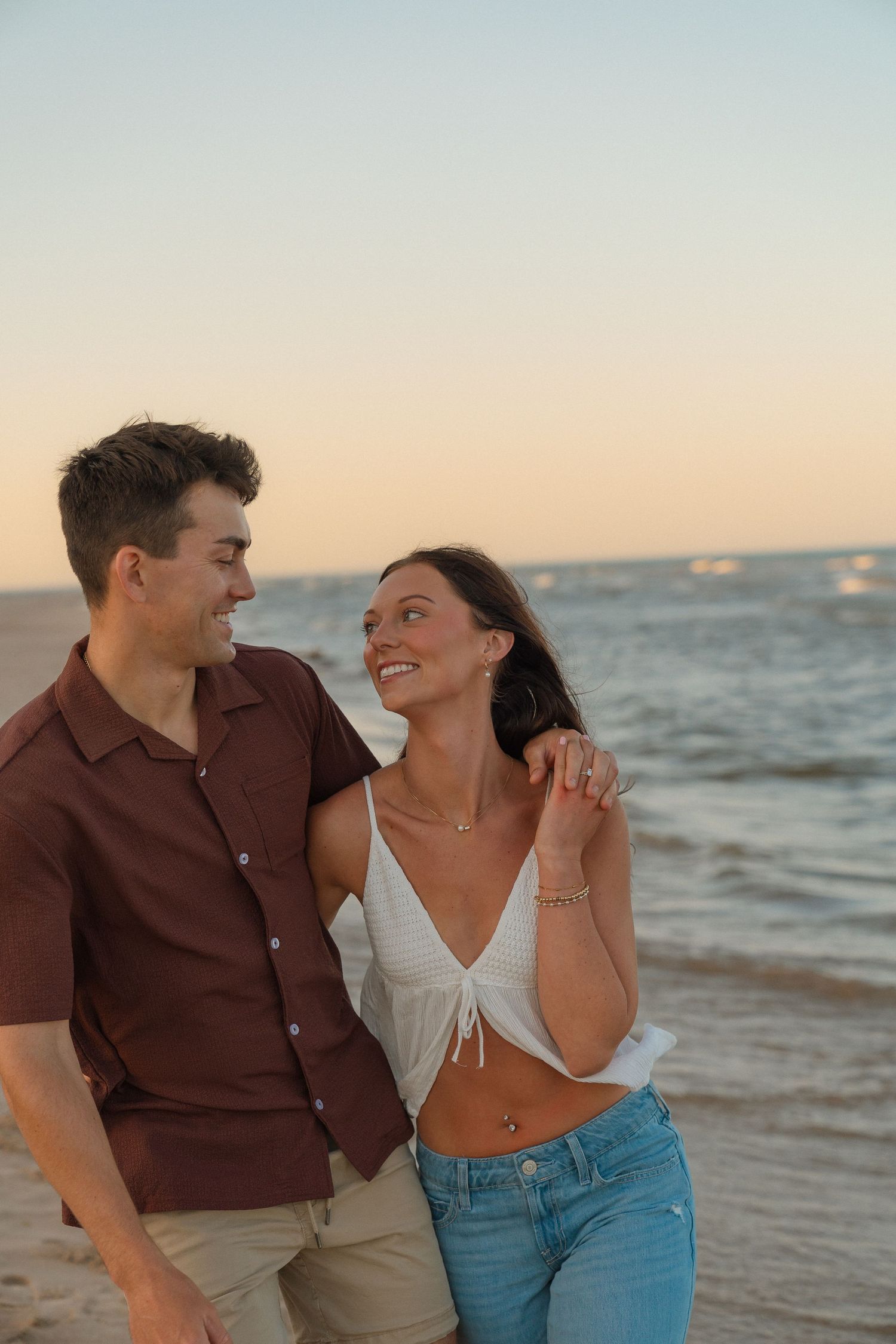 A young couple standing together on a beach at sunset, looking affectionately at each other.