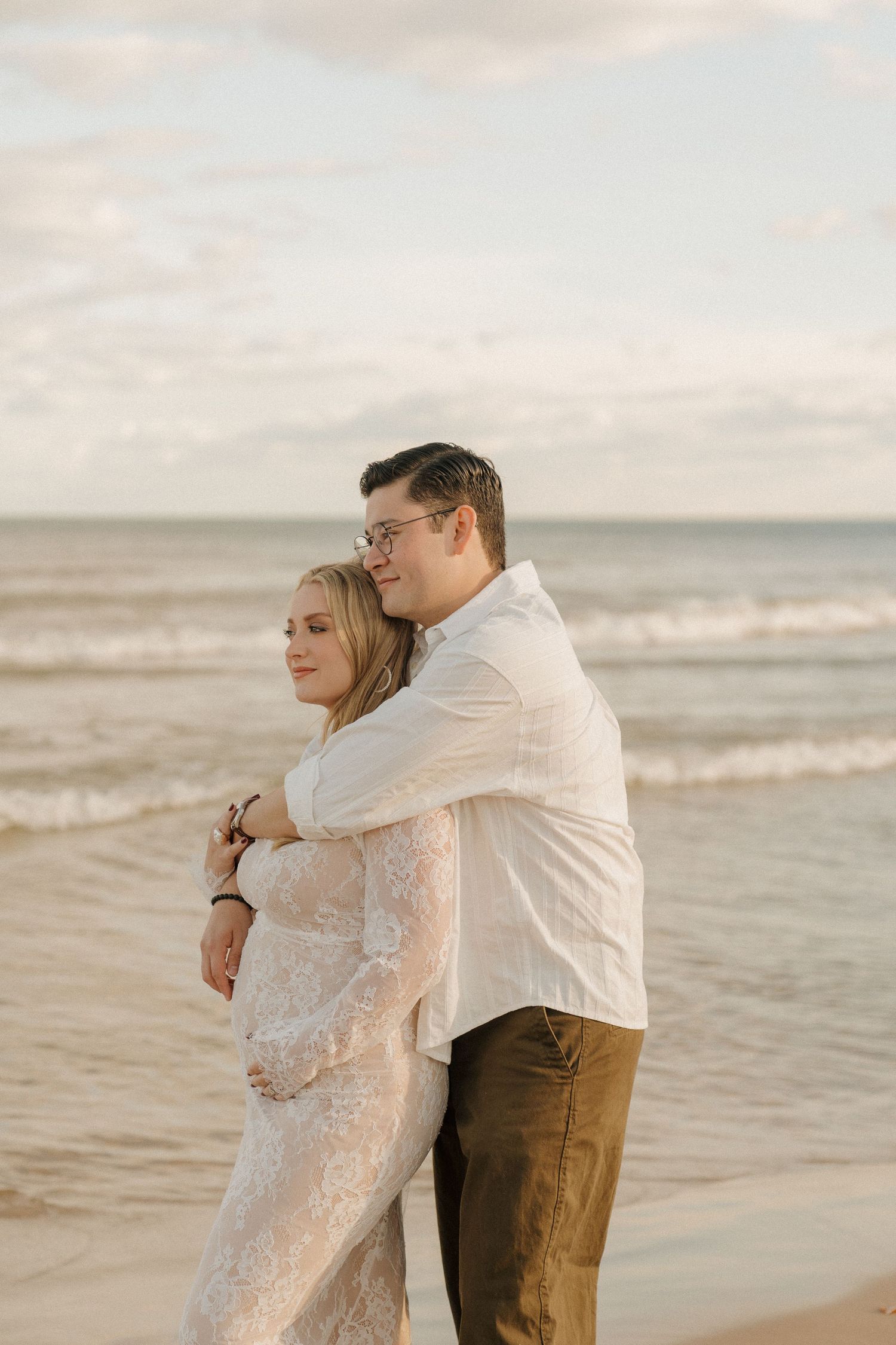 Man tenderly embracing pregnant partner in white lace dress on beach with ocean waves behind them.