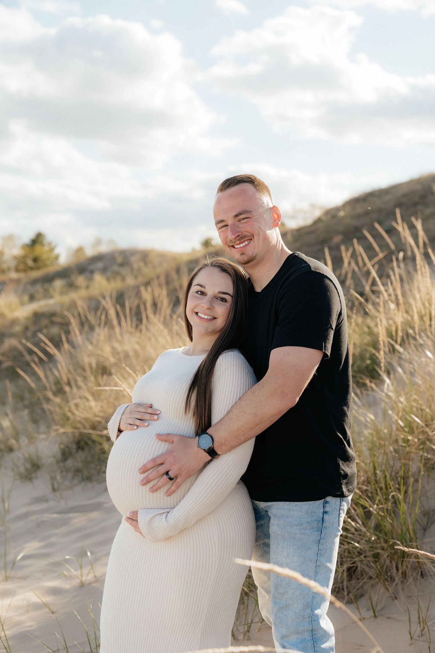 Couple posing on sandy dunes, woman in white dress cradling pregnant belly, man in black shirt embracing her from behind.