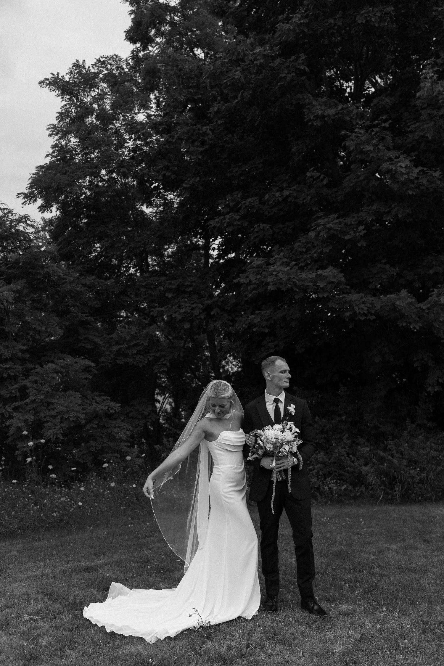 Black and white photo of a bride in a flowing veil and groom in a suit holding a bouquet, standing on a lawn.