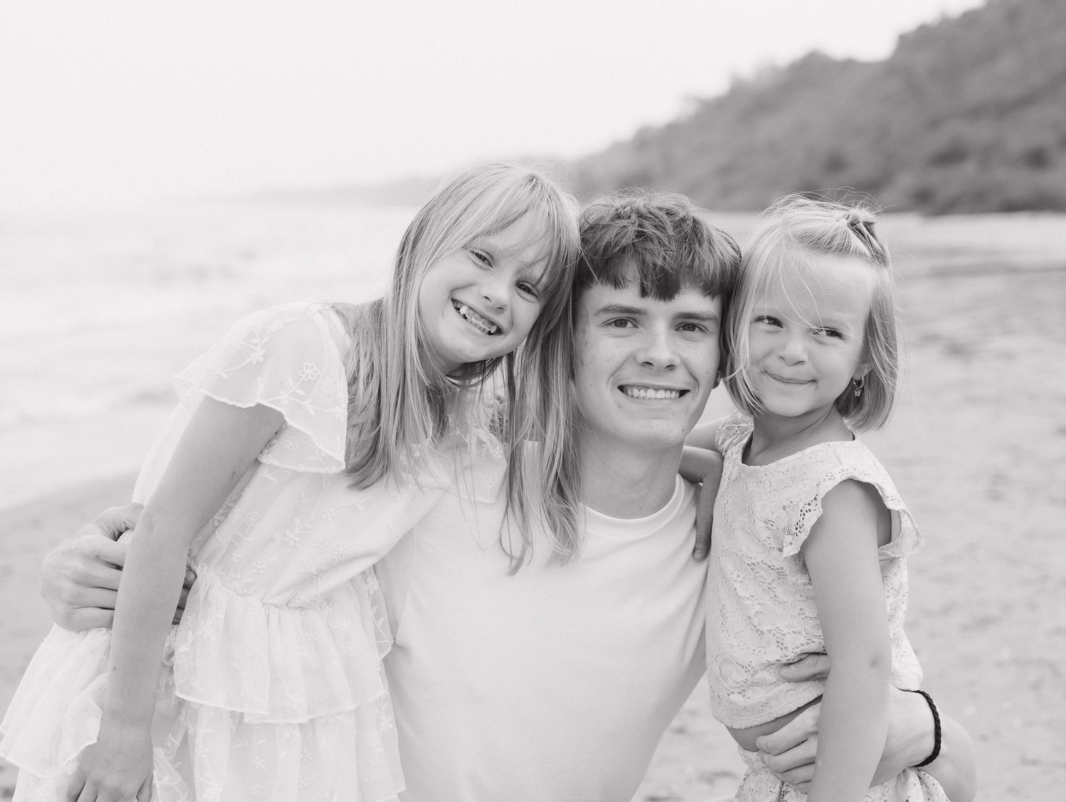 A black and white image shows a happy family of three - a brother and two young daughters - smiling together on a beach.
