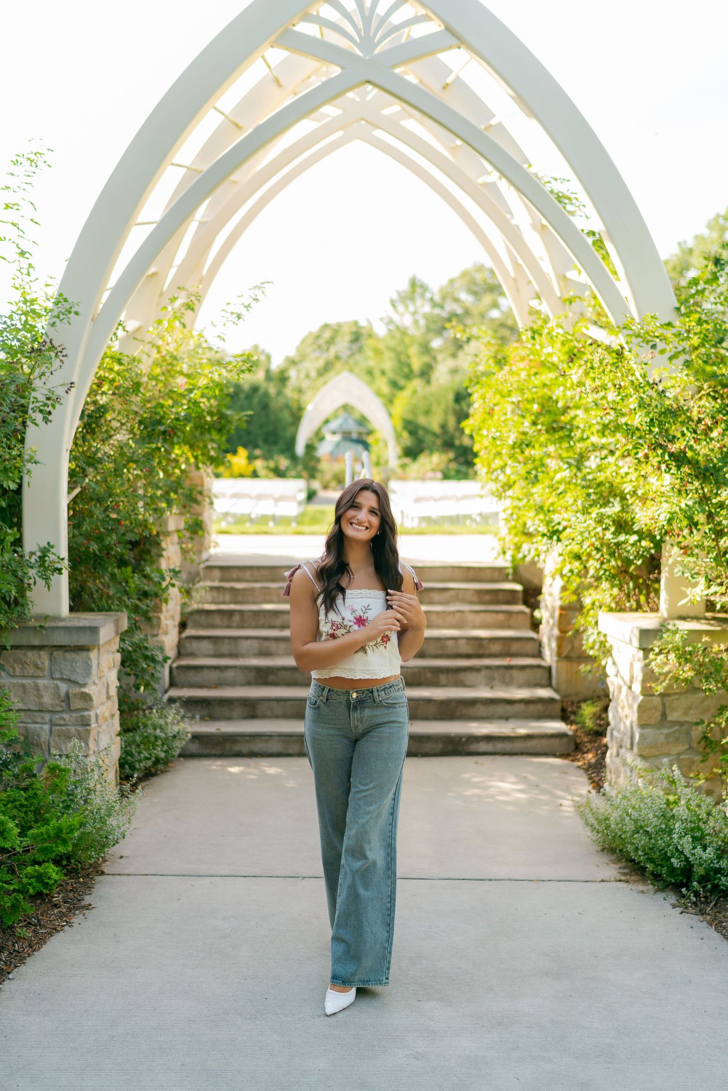 Young woman in wide-leg jeans and floral top smiling under white gothic arch with lush greenery and stone steps.