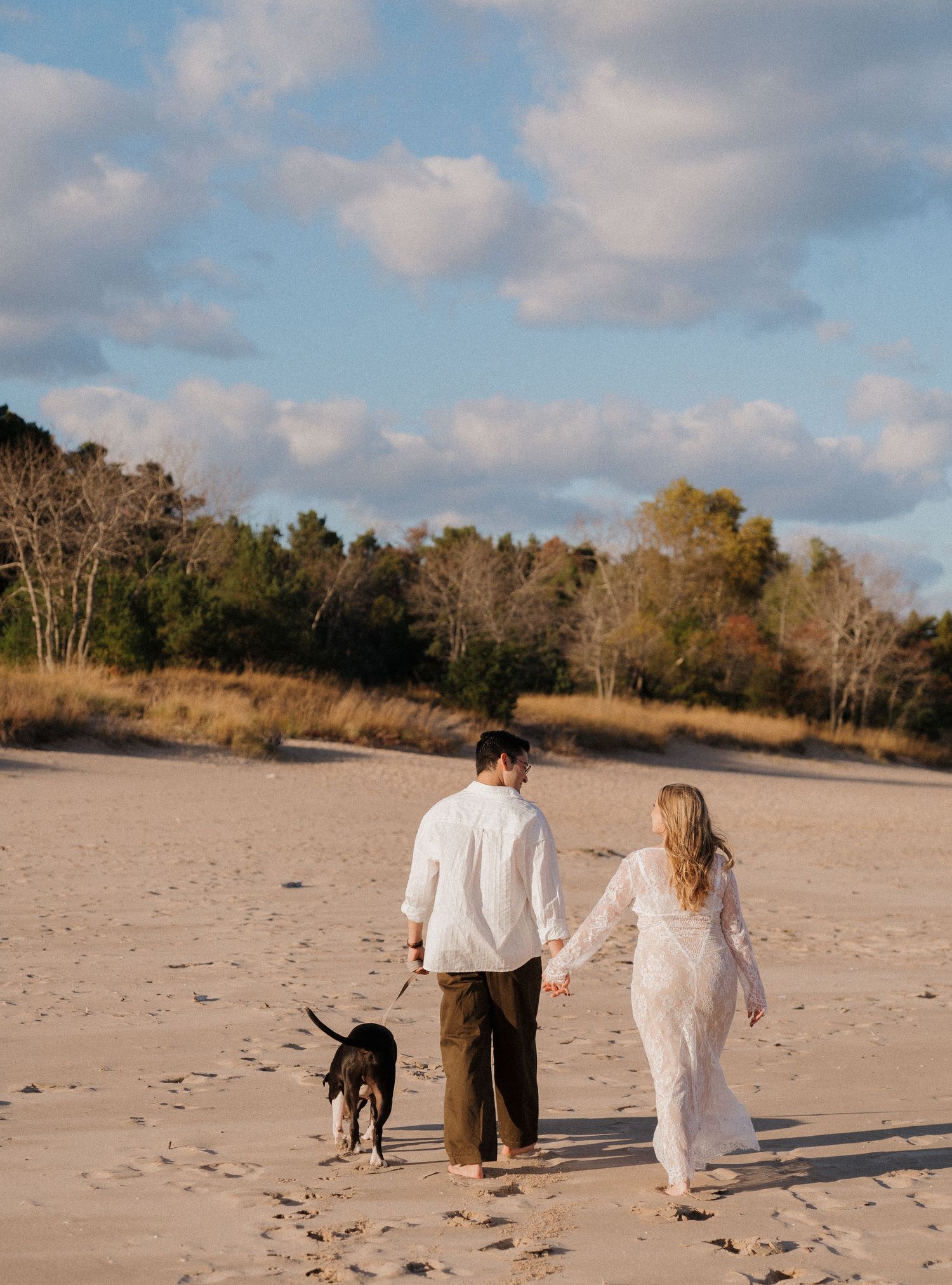 Couple walking dog on sandy beach, woman in lace dress, man in white shirt, trees in background.