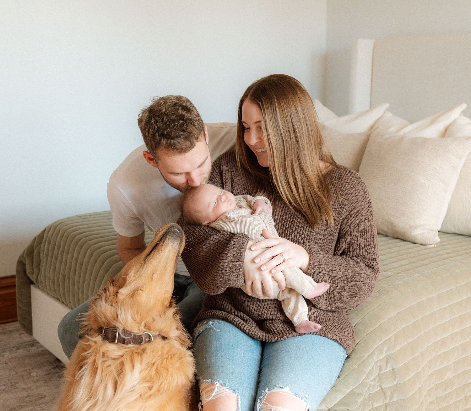 A couple sits on a couch with a dog, cuddling a newborn baby and enjoying a tender family moment.