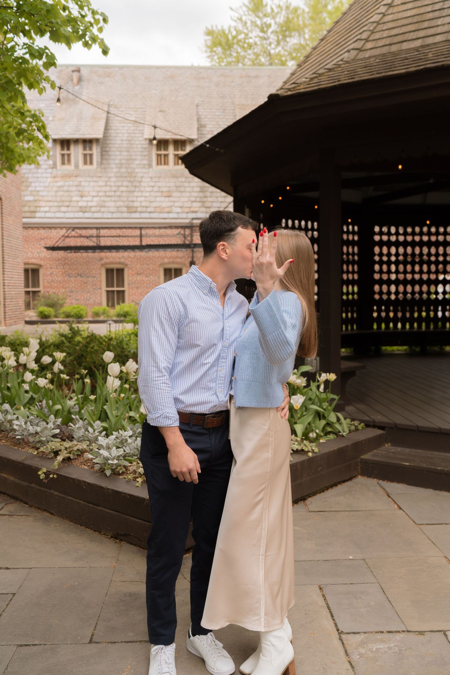 A young couple embracing in a garden setting with a traditional building in the background.