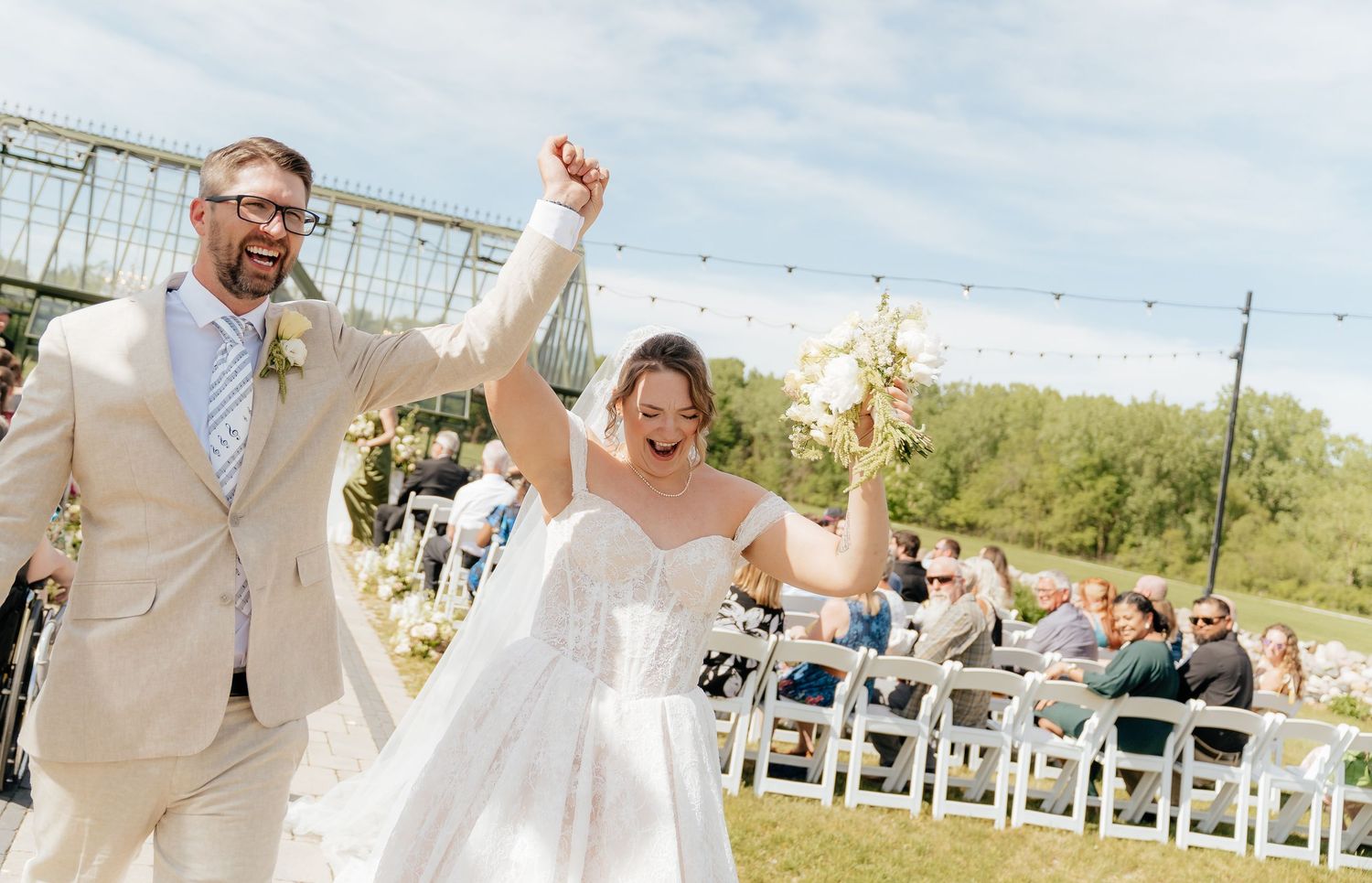 Joyful newlyweds raise hands walking down outdoor wedding aisle, bride in white gown, groom in tan suit, guests seated.
