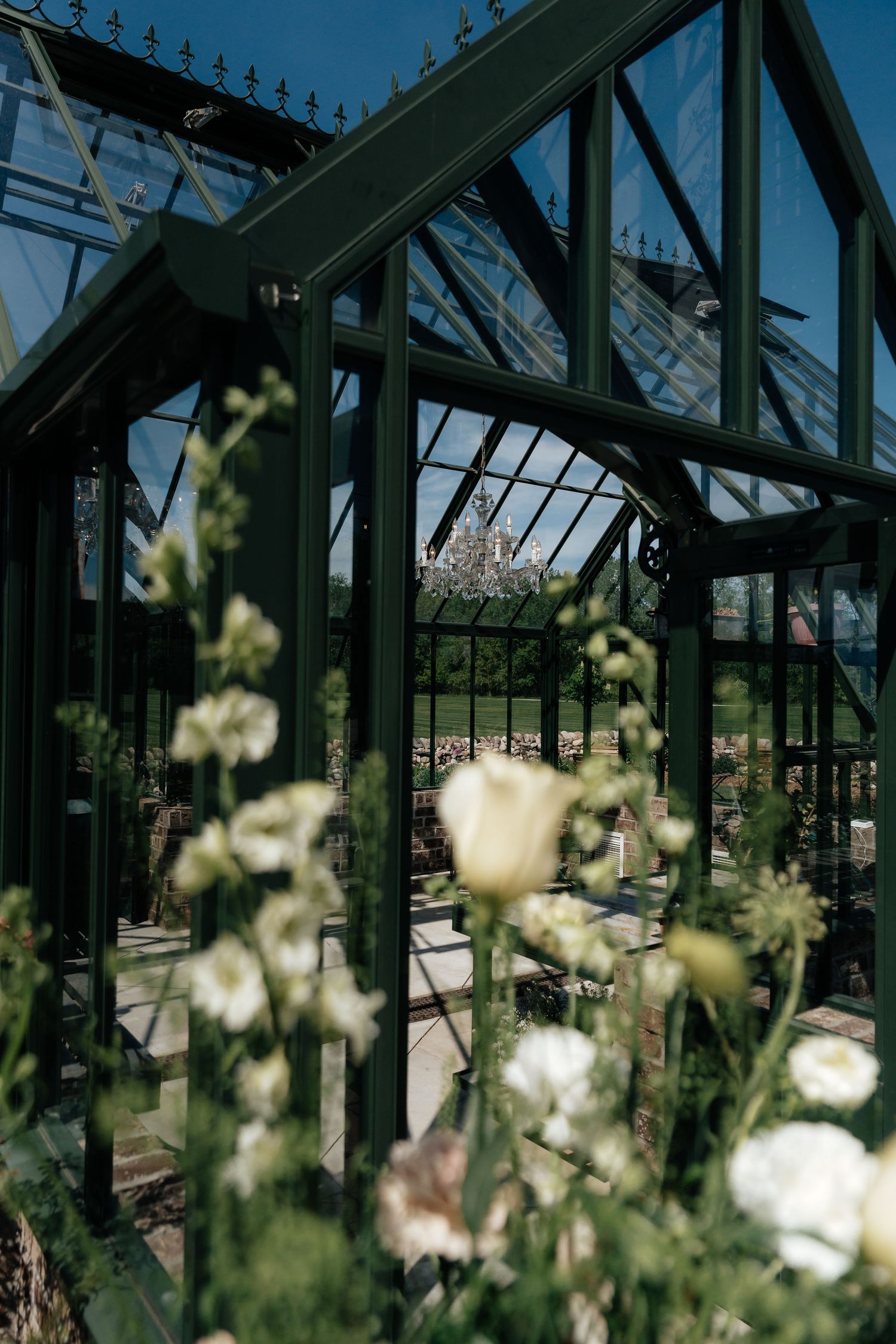 Green glass greenhouse with white flowers in foreground under blue sky, elegant Victorian-style botanical garden structure.