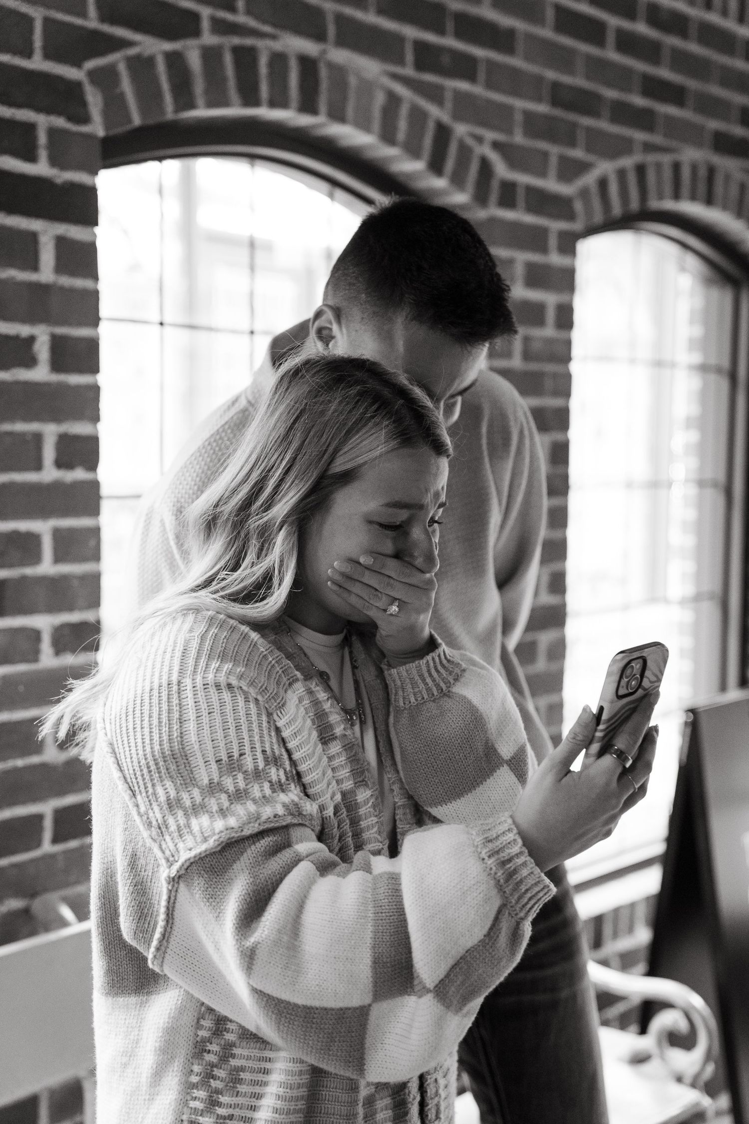 Black and white photo of a couple sharing an emotional moment, woman covering her mouth in surprise near brick wall.
