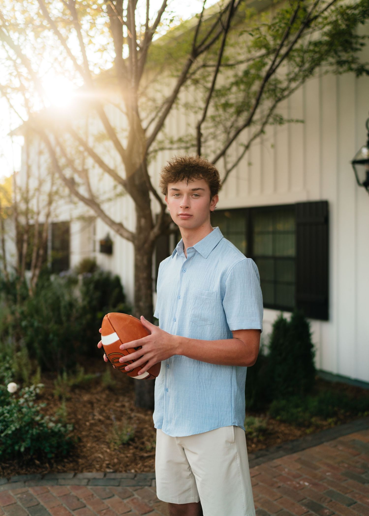 Teen boy holding a football in a light blue shirt and khaki shorts, posing outdoors at golden hour.