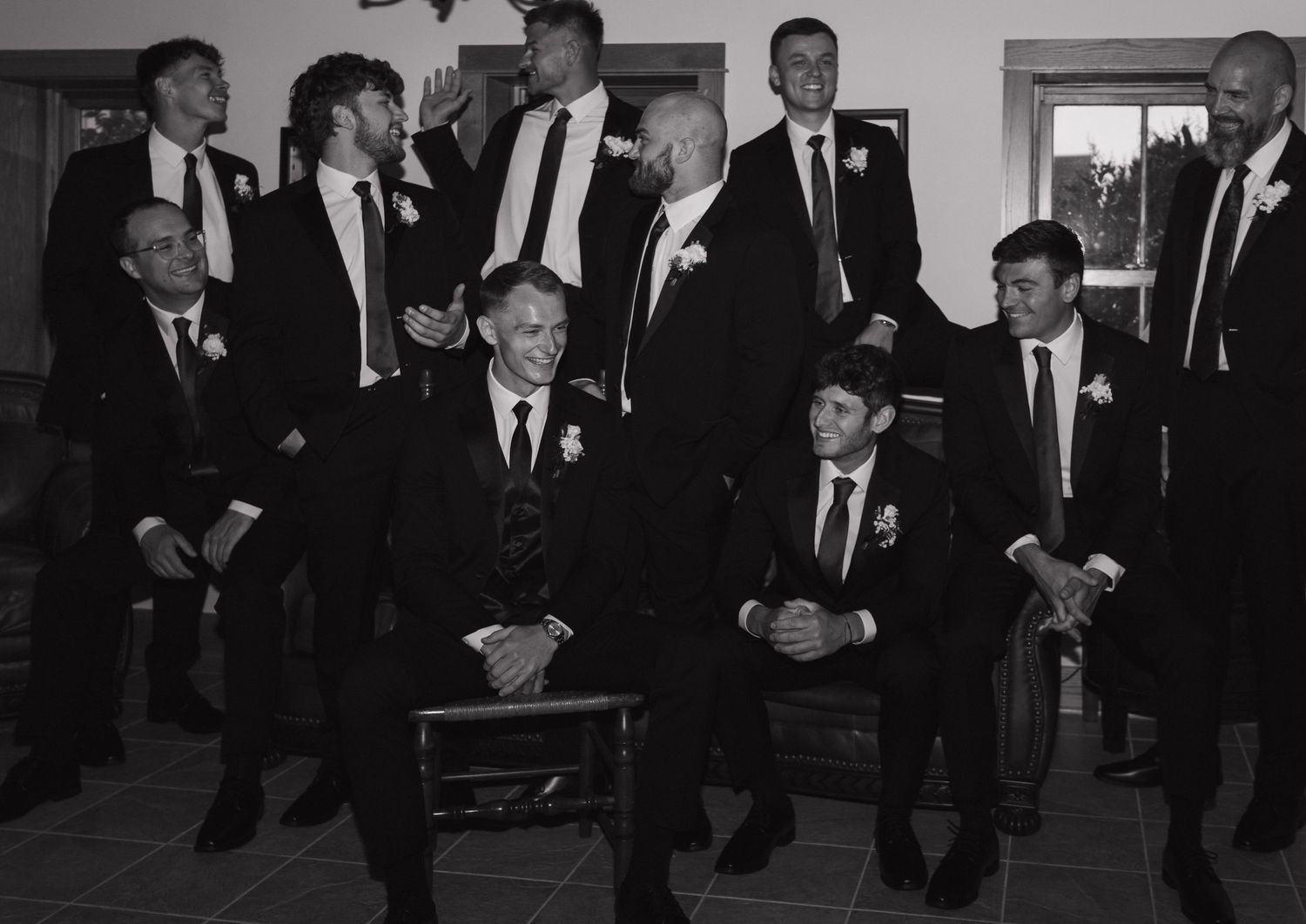 Black and white photo of a large group of men in formal suits with boutonnieres, posing together at a wedding.