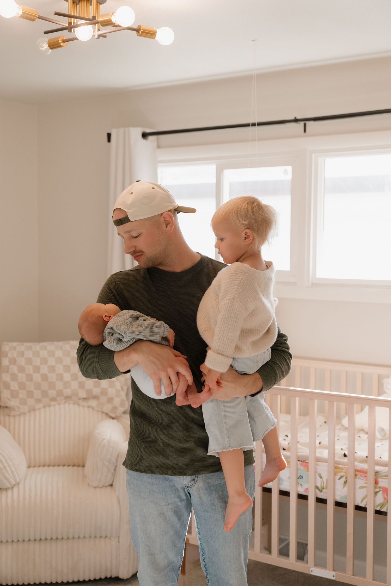 A man in a black shirt is holding a young child in a cozy nursery with a chandelier overhead.