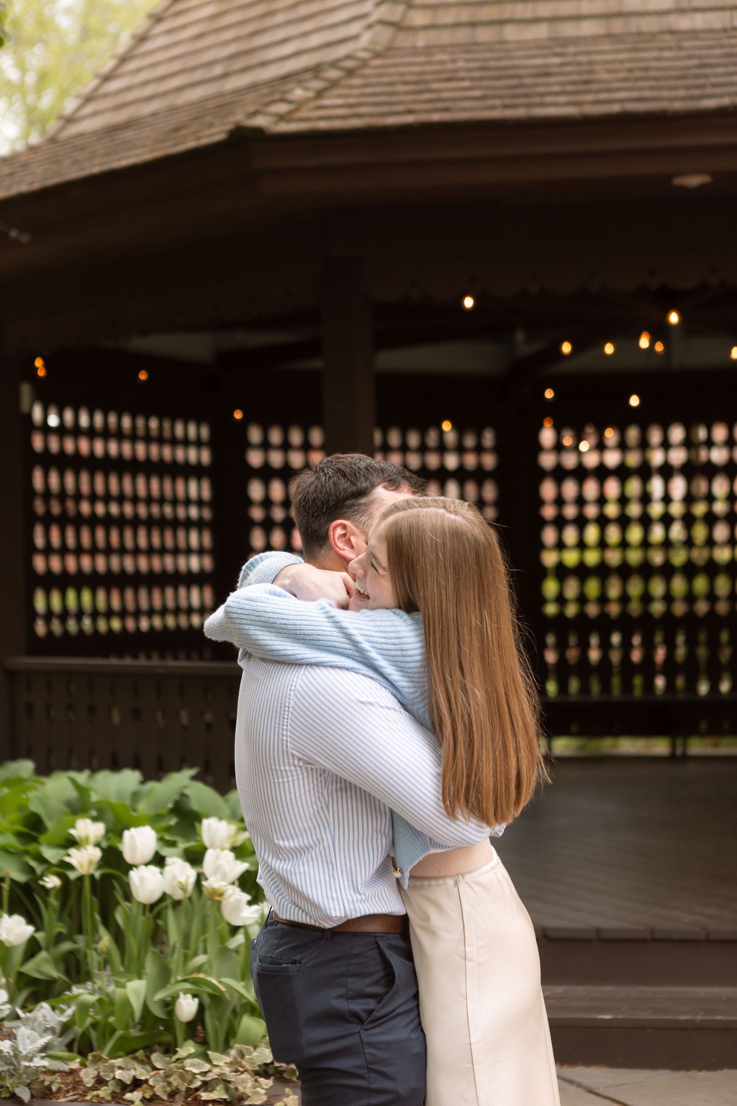 A couple embracing in front of a wooden gazebo with string lights, surrounded by greenery.