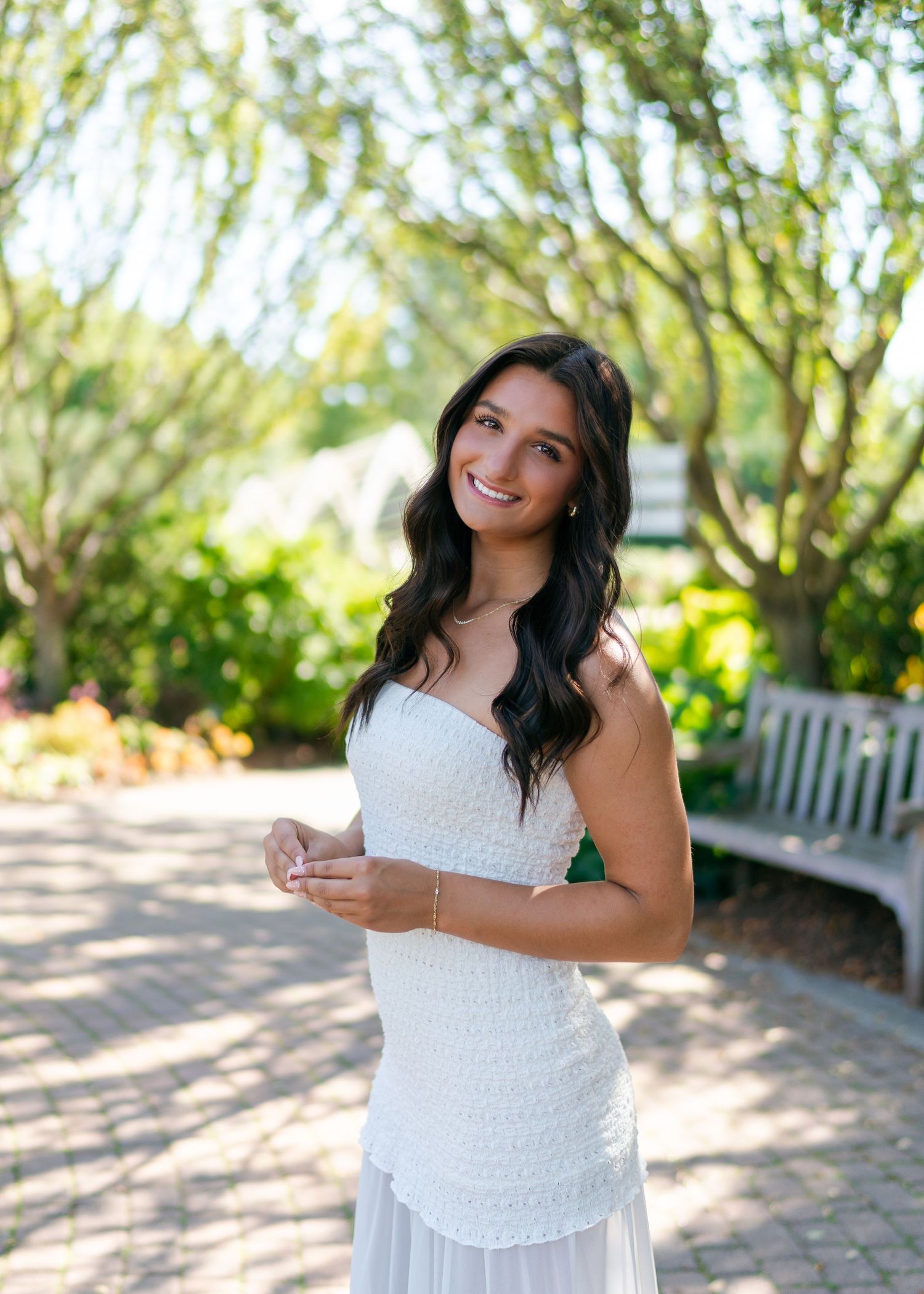 Young woman in white dress smiling in a garden with lush greenery and a bench backdrop.