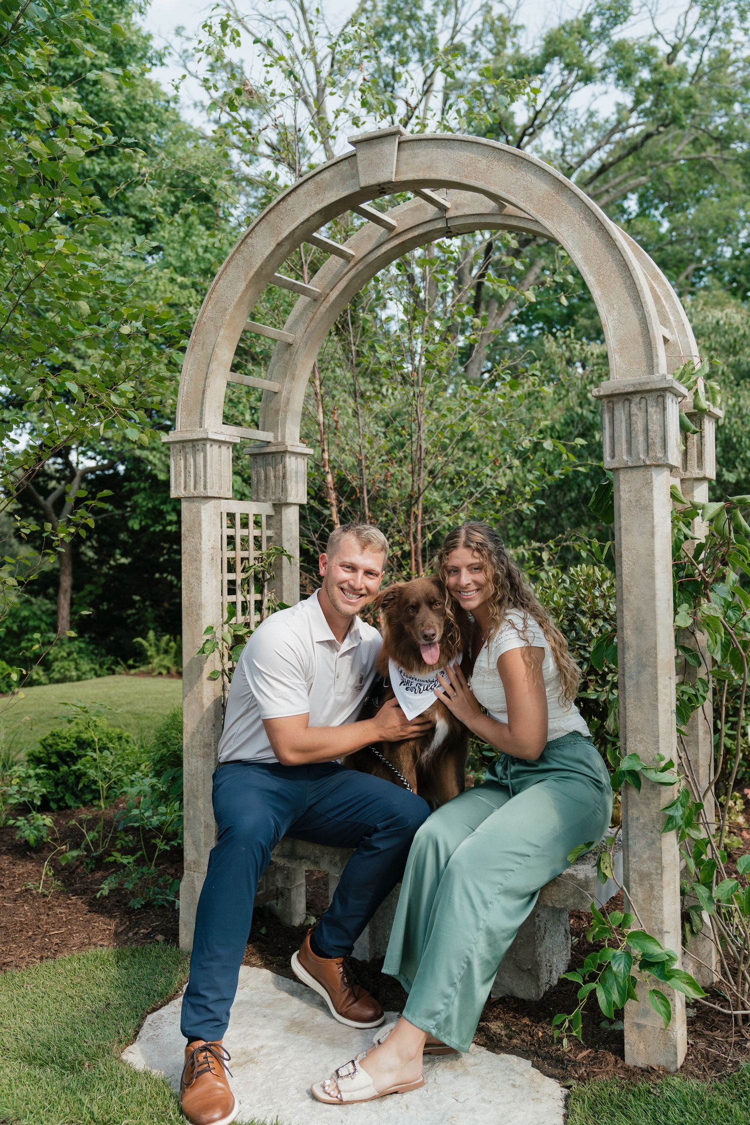 Couple sitting under stone garden arch with brown dog wearing bandana, surrounded by lush greenery.