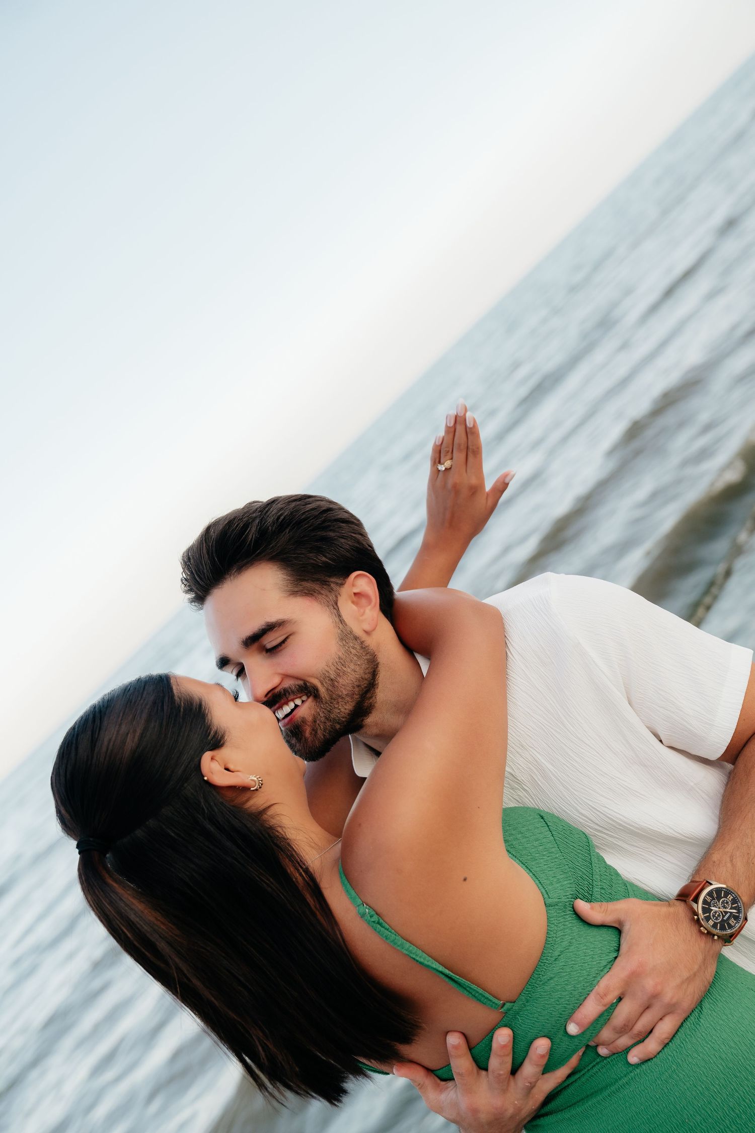Romantic couple embracing and almost kissing by the ocean, woman in green dress, man in white shirt wearing a watch.