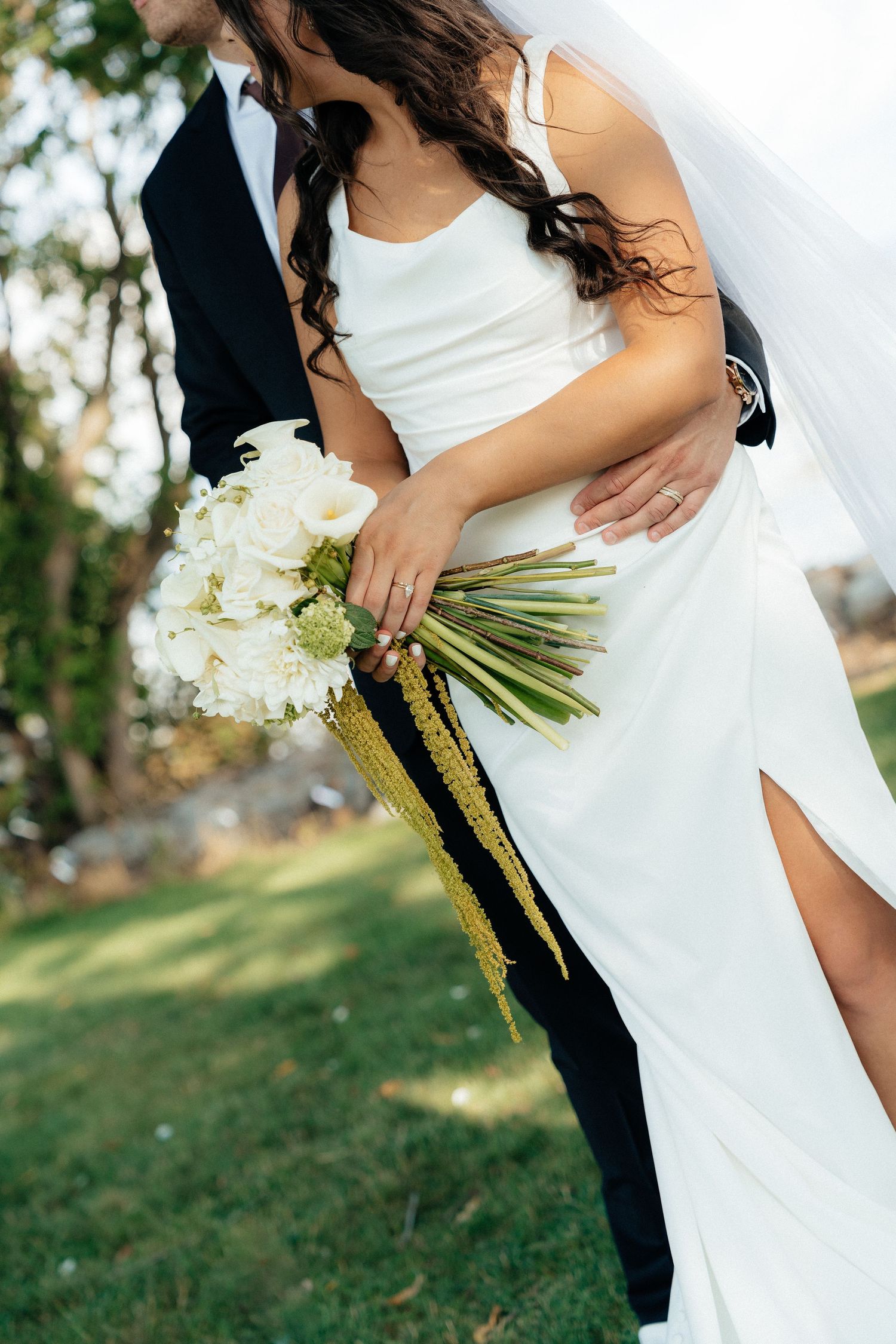 Bride in white gown holding floral bouquet embraced by groom in black suit outdoors on green lawn.