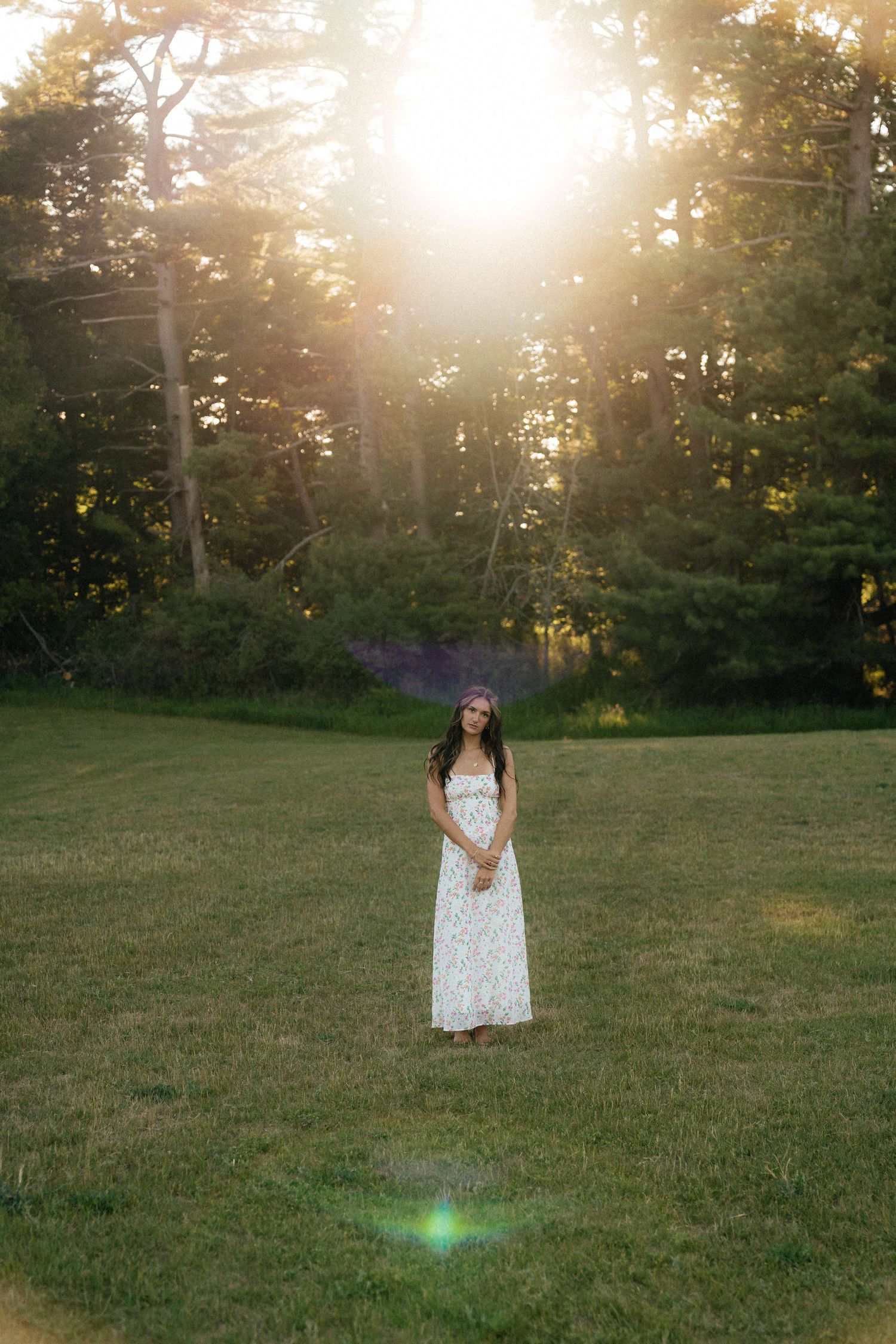 Young woman in white floral maxi dress standing in sunlit green meadow surrounded by tall trees at golden hour.