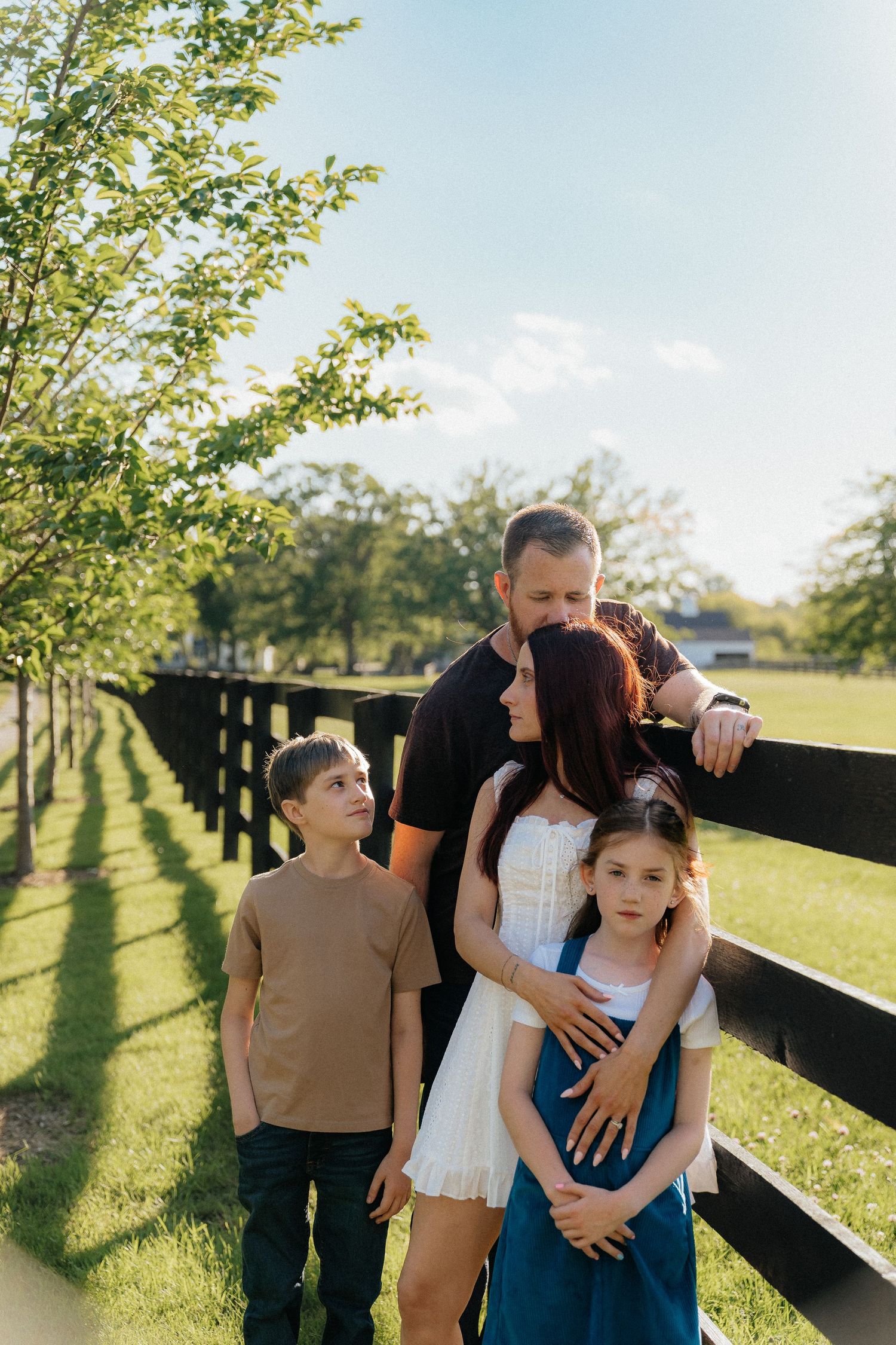 Family of four posing by a wooden fence in a sunny green park, parents embracing two children on a bright summer day.