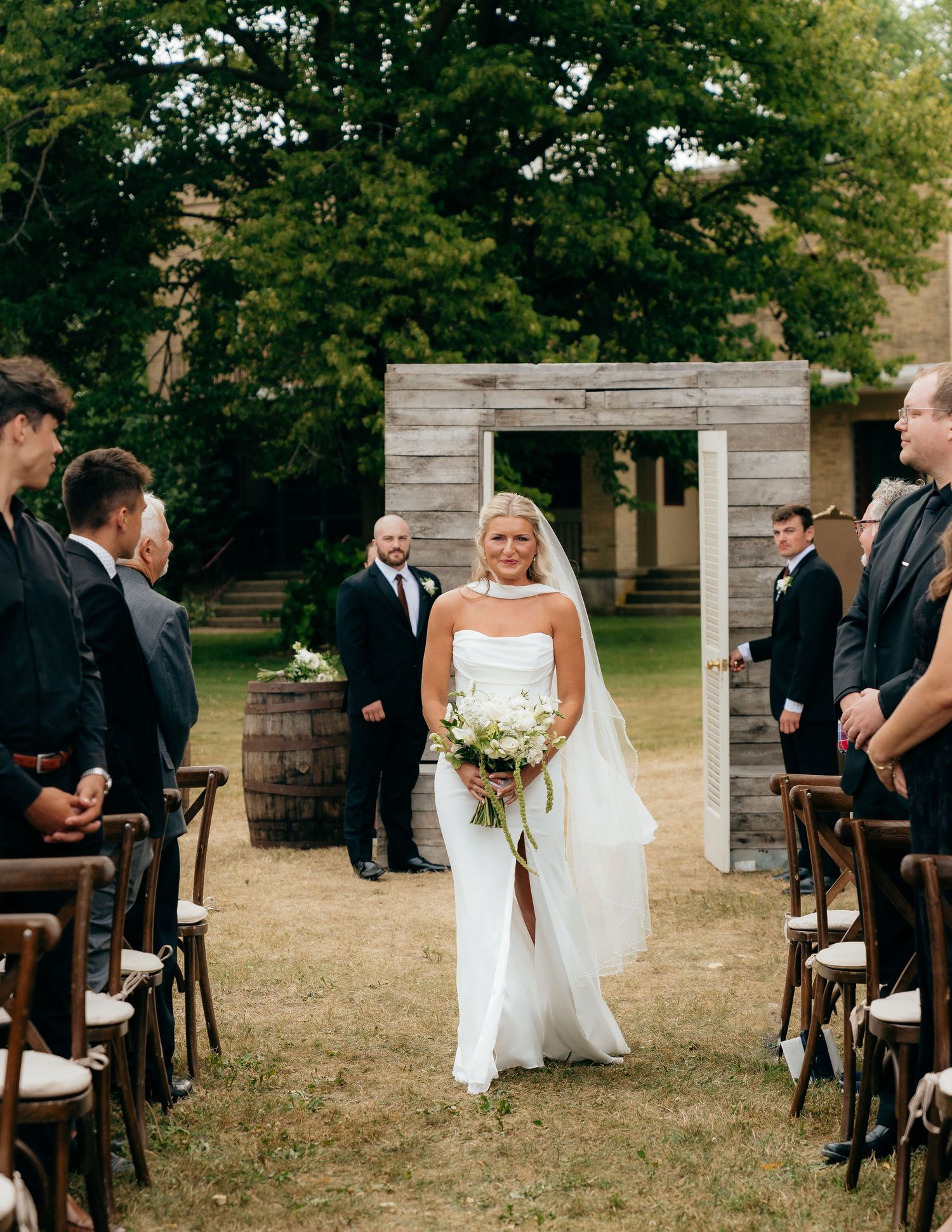 Bride walks down outdoor aisle in white gown and veil, holding bouquet, with guests and rustic wooden arch backdrop.