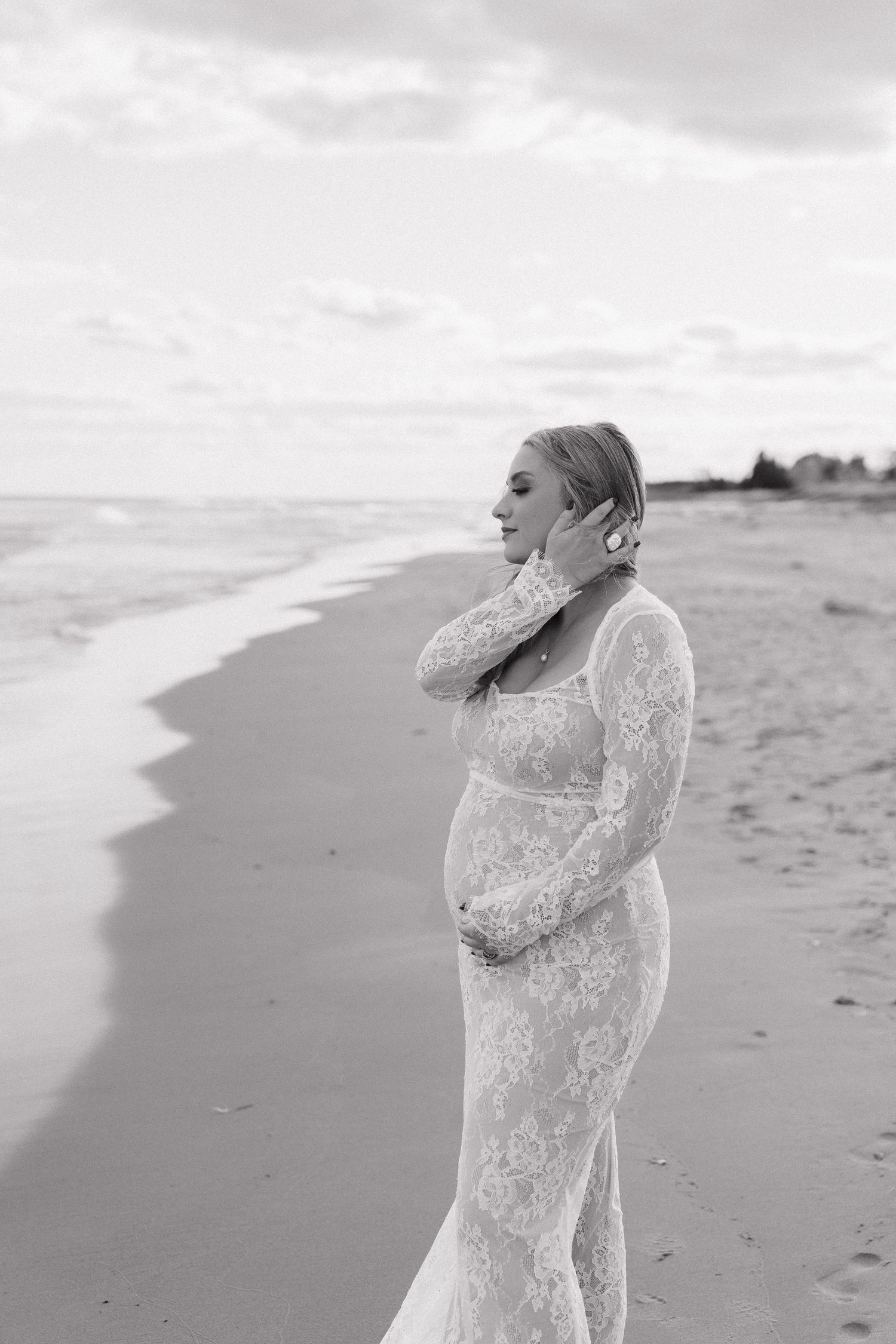 Pregnant woman in lace gown poses on beach shoreline in elegant black and white maternity photo.