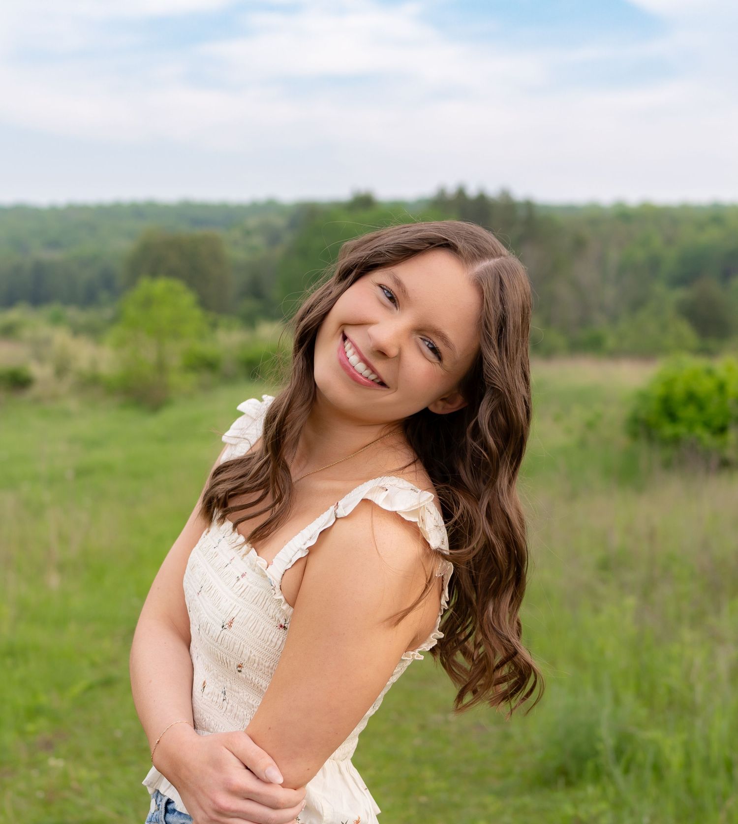 A smiling young woman with long wavy brown hair stands in a grassy field with trees in the background.