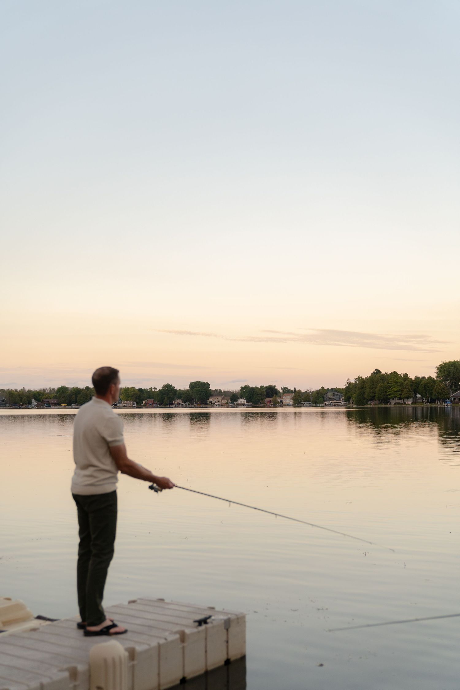 A person fishing on a wooden dock at a serene lake during a colorful sunset.