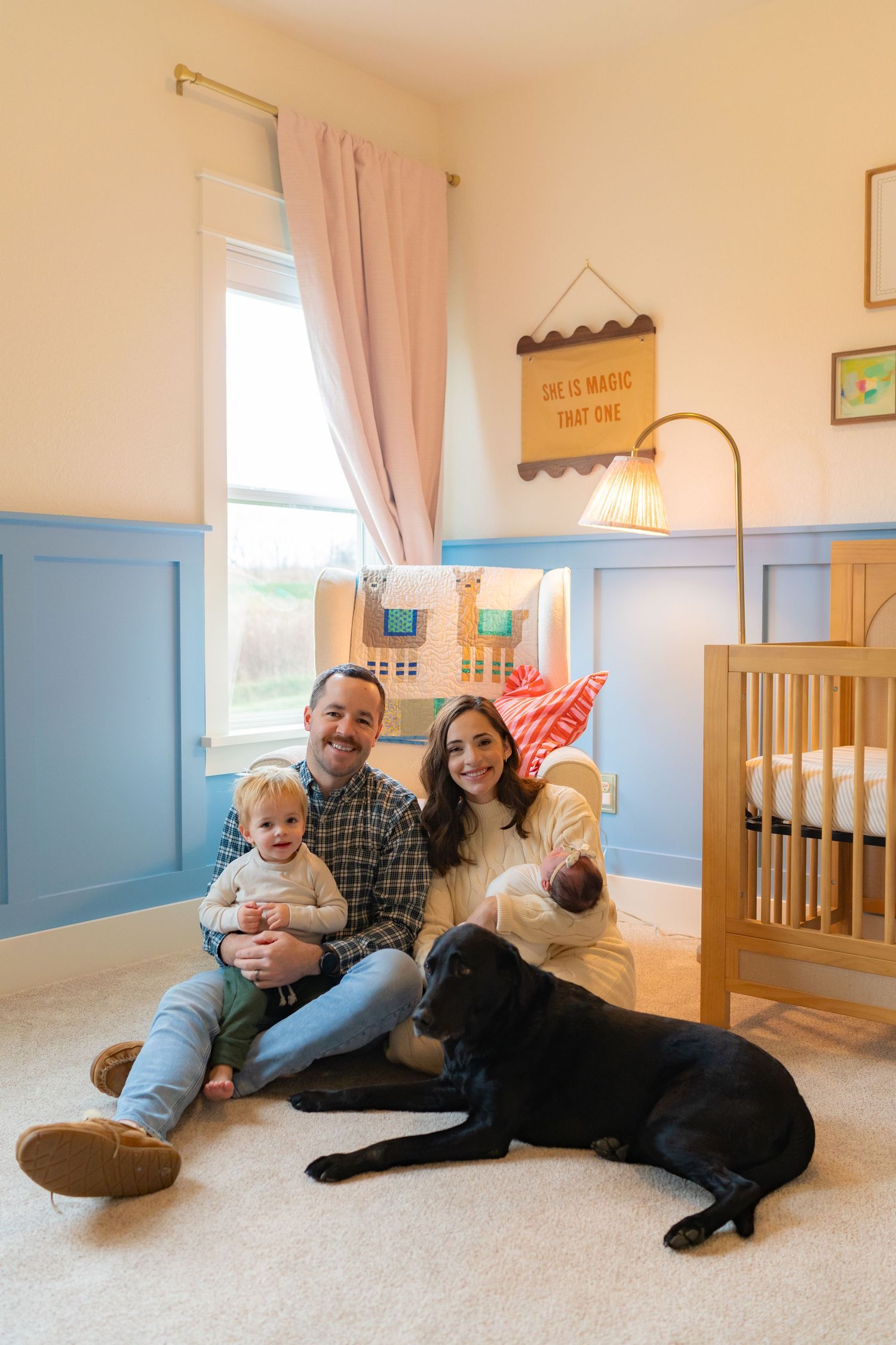 A family of three, including a young child, sitting on the floor with their dog in a cozy nursery room.