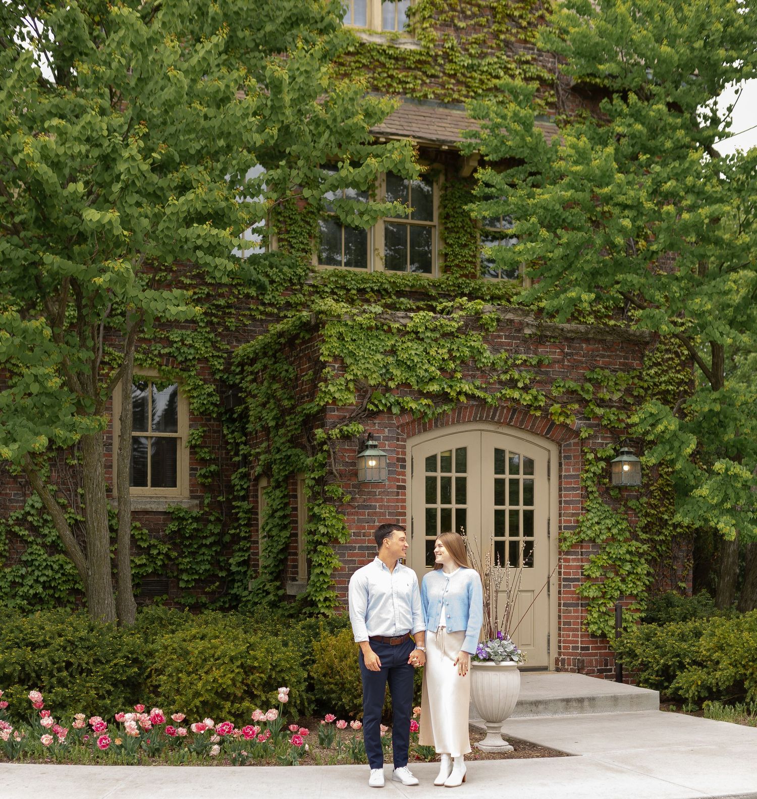 A couple standing in front of a historic building surrounded by lush greenery and flowers.