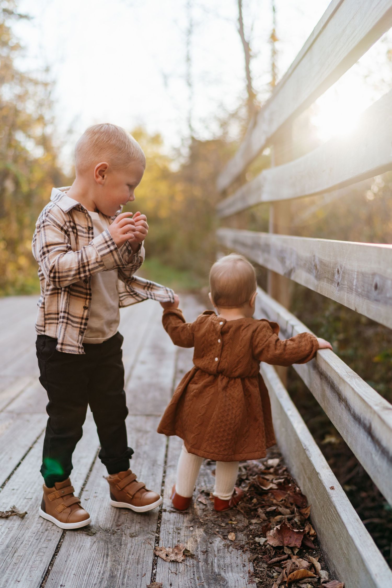 Two young children holding hands on a wooden bridge surrounded by autumn foliage in golden sunlight.