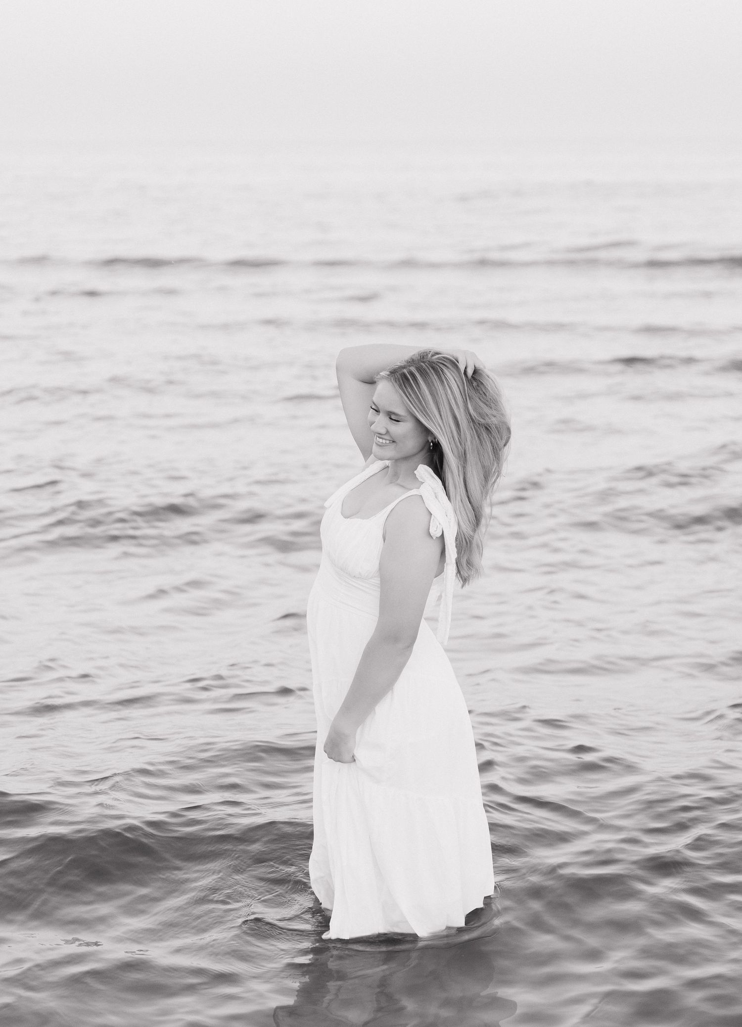 A woman in a white dress standing in the ocean, with waves in the background.