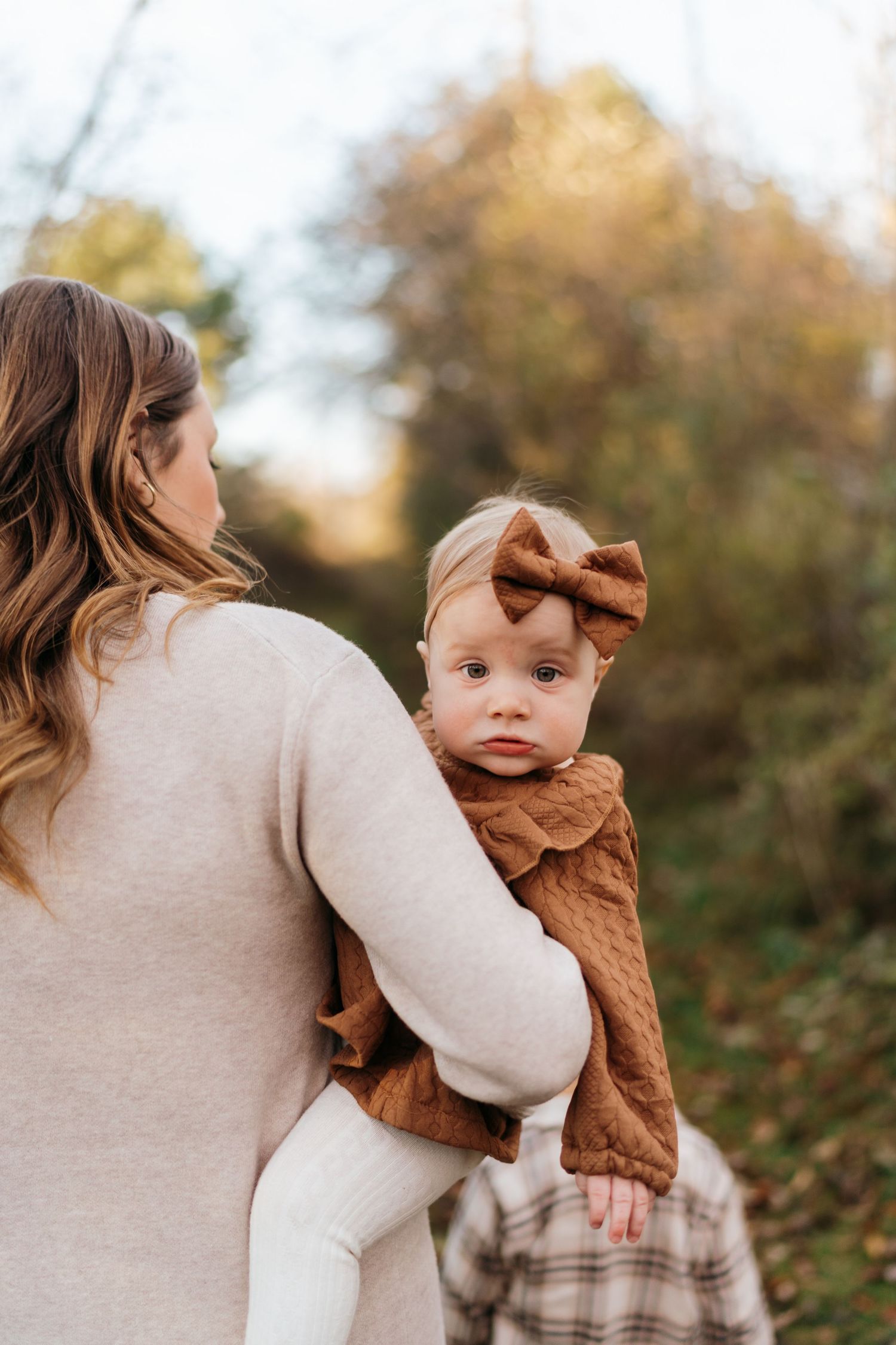 A mother holds her baby girl in a brown bow headband and rust dress outdoors in autumn foliage.