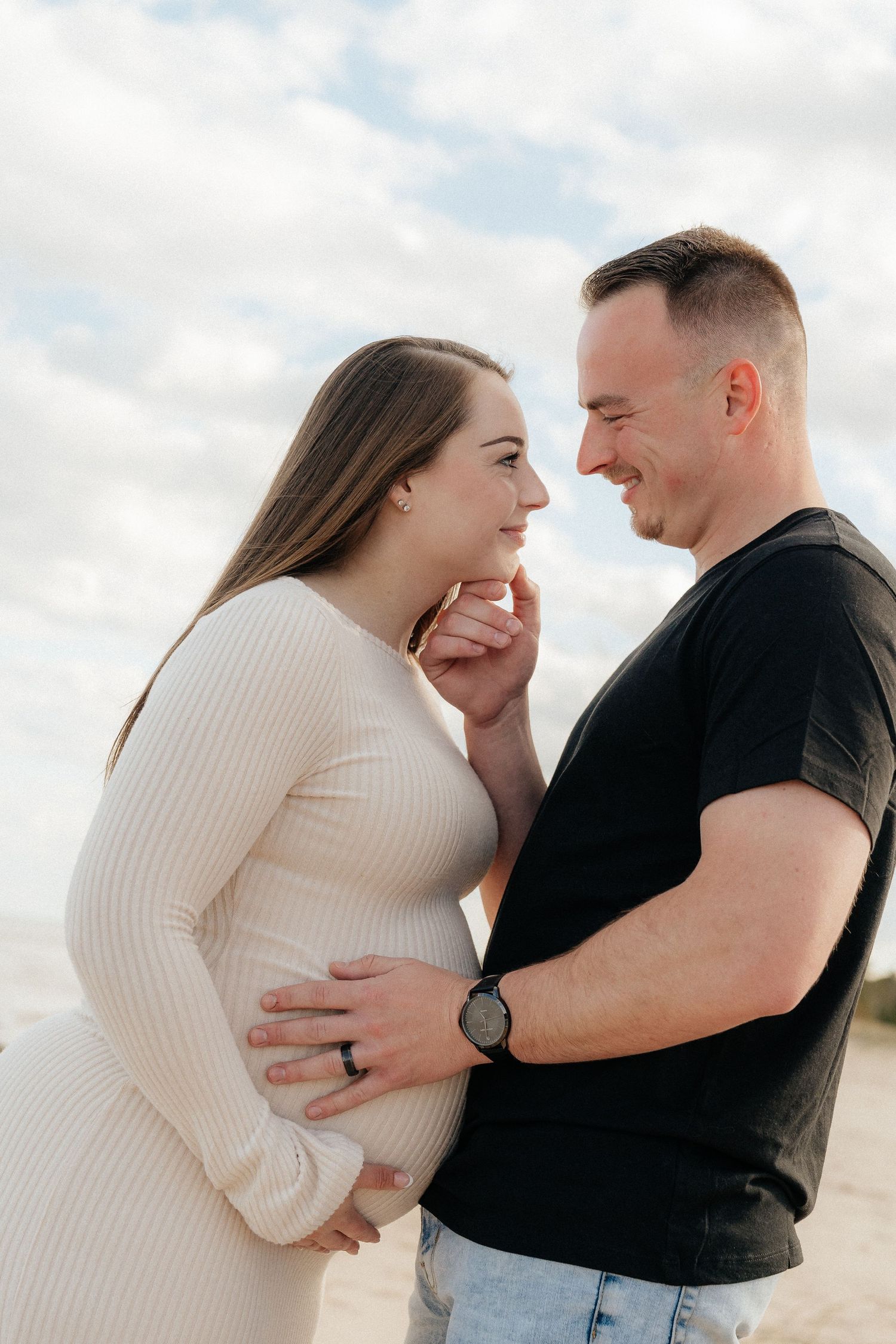A smiling couple shares a tender moment on a sandy beach, with the man gently holding his pregnant partners belly.
