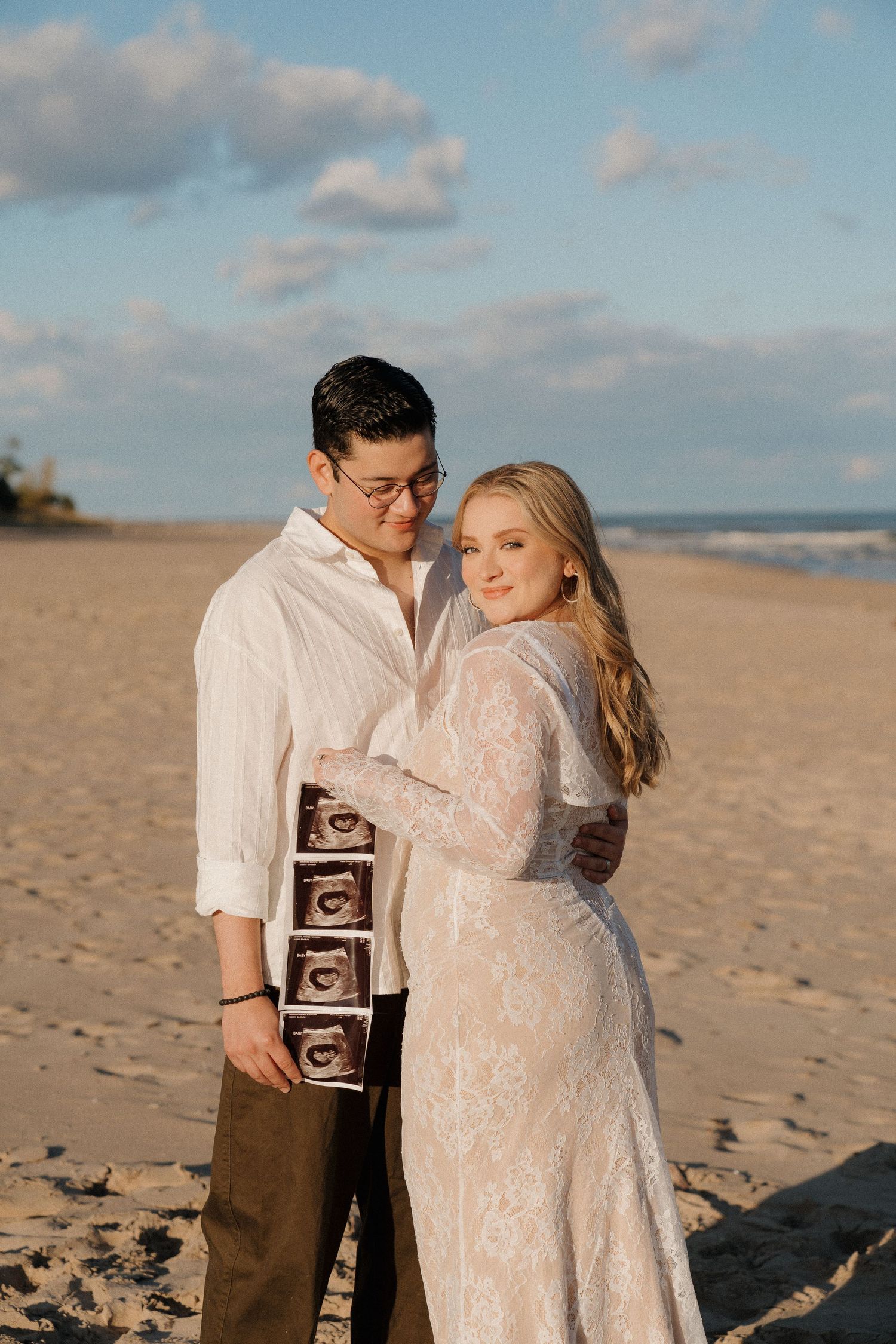 Couple holding ultrasound photos on sandy beach, woman in white lace maternity dress at sunset.