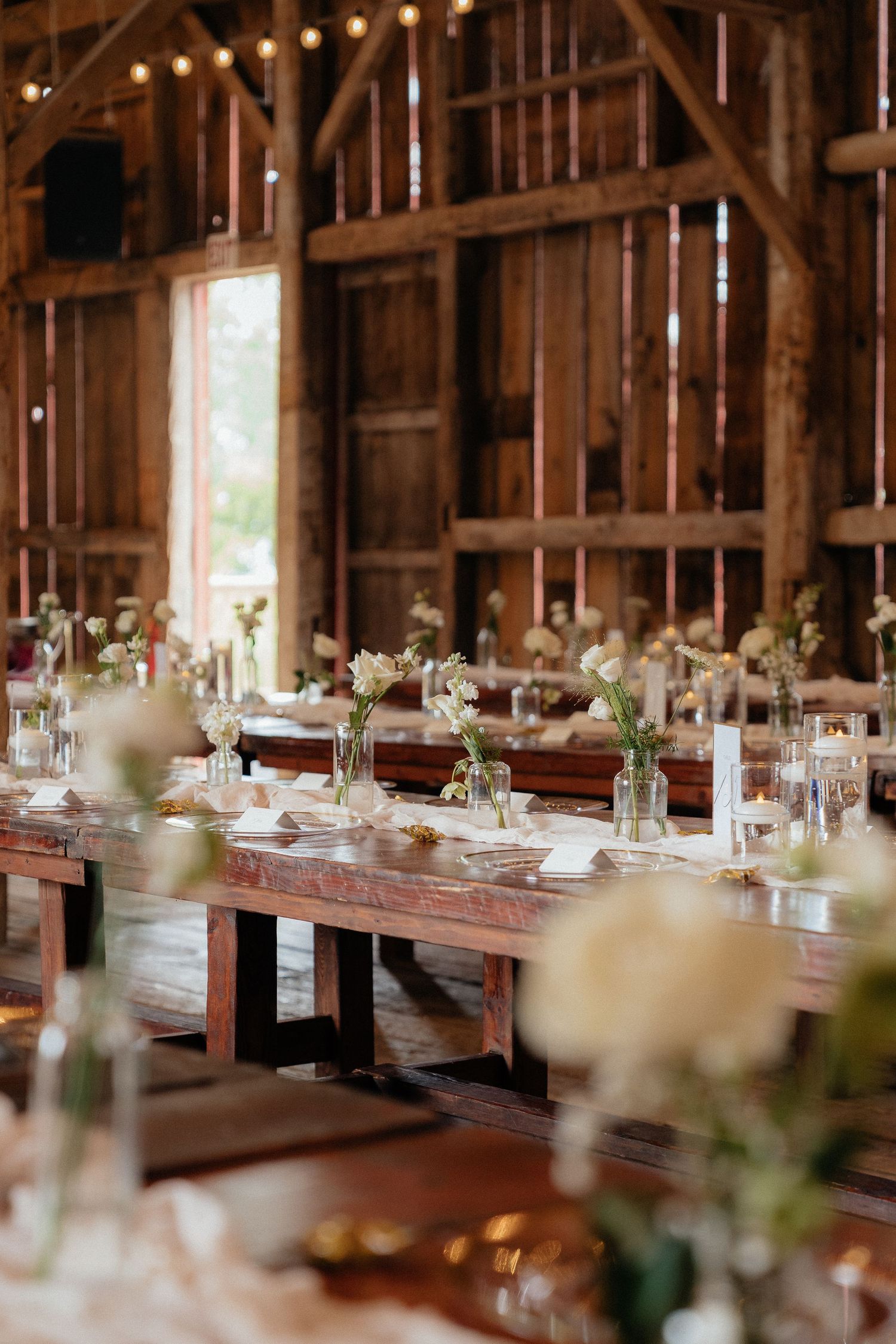 Rustic barn wedding reception with long wooden tables, white floral centerpieces, candles, and string lights overhead.