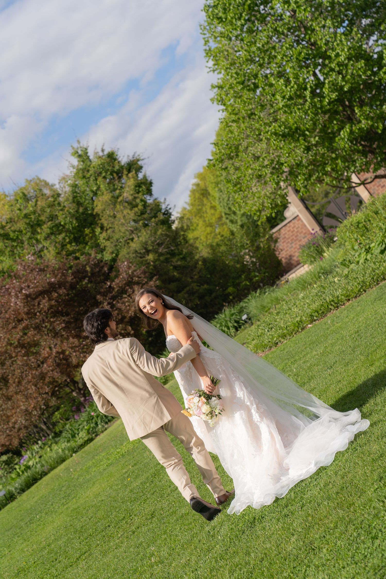 A bride and groom walking on a grassy hill surrounded by trees and a blue sky with clouds.