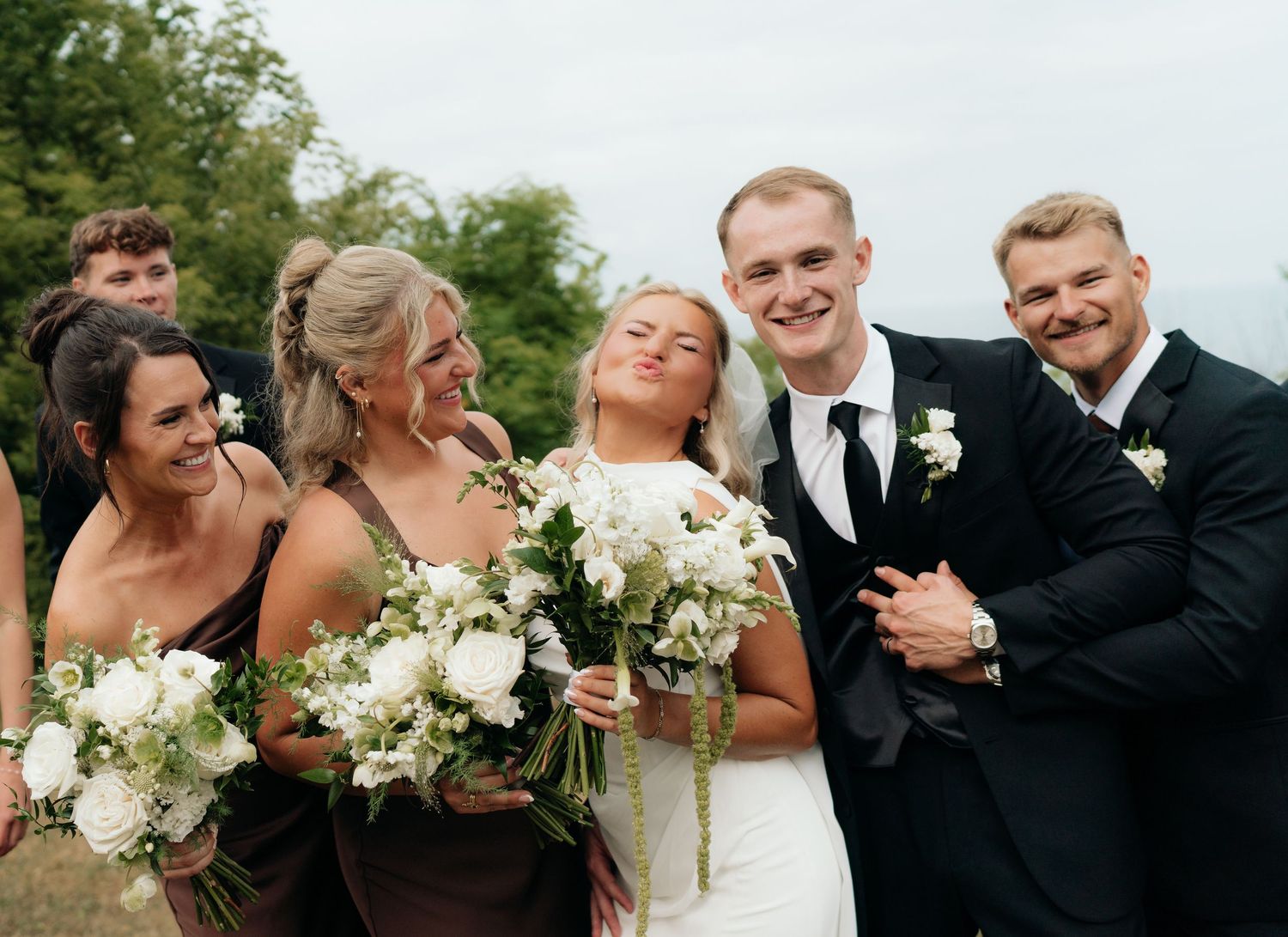 Wedding party smiling outdoors, bride in white gown holding bouquet, bridesmaids in brown dresses, groomsmen in black suits.