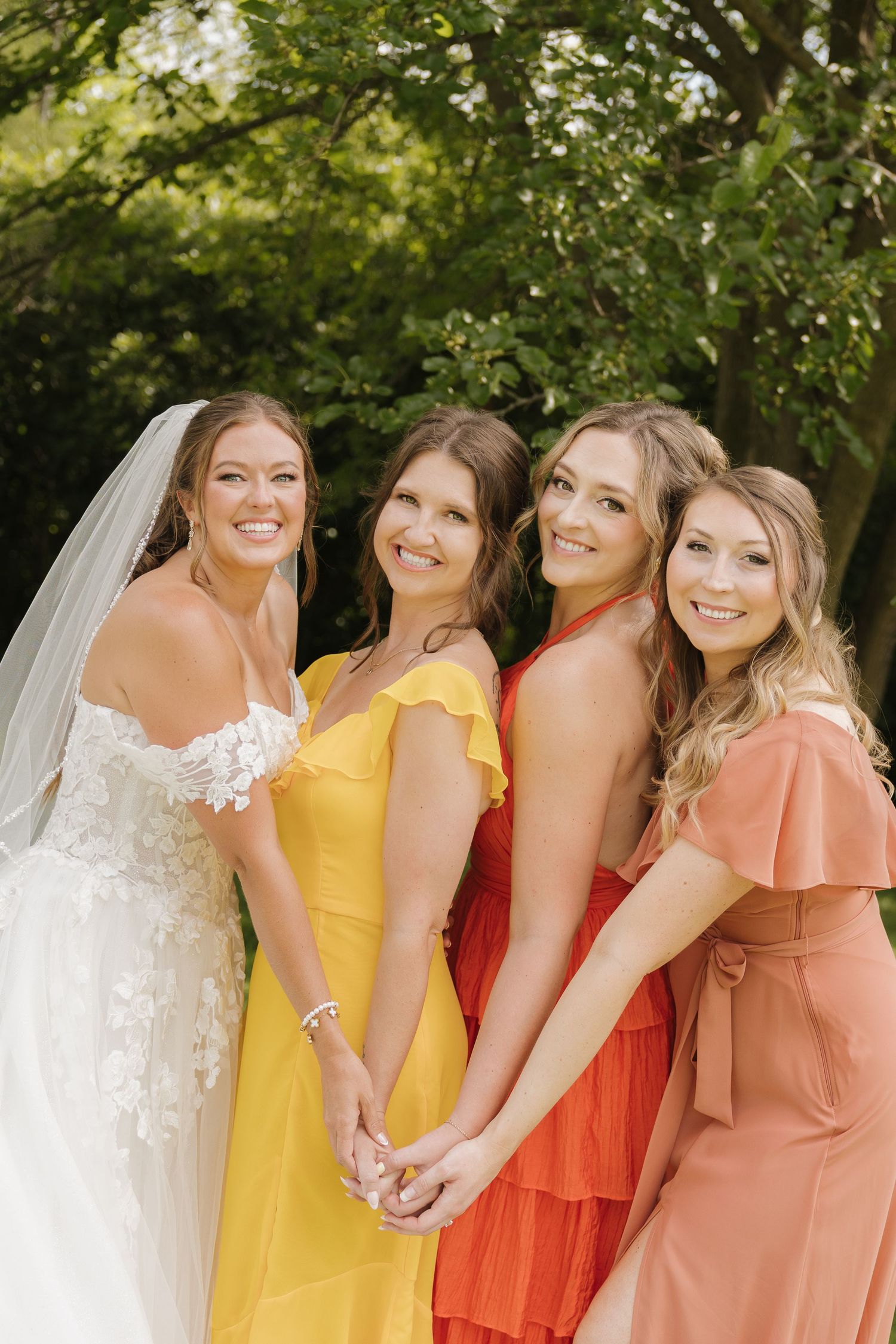 A group of four smiling women in colorful dresses stand together in a garden setting.
